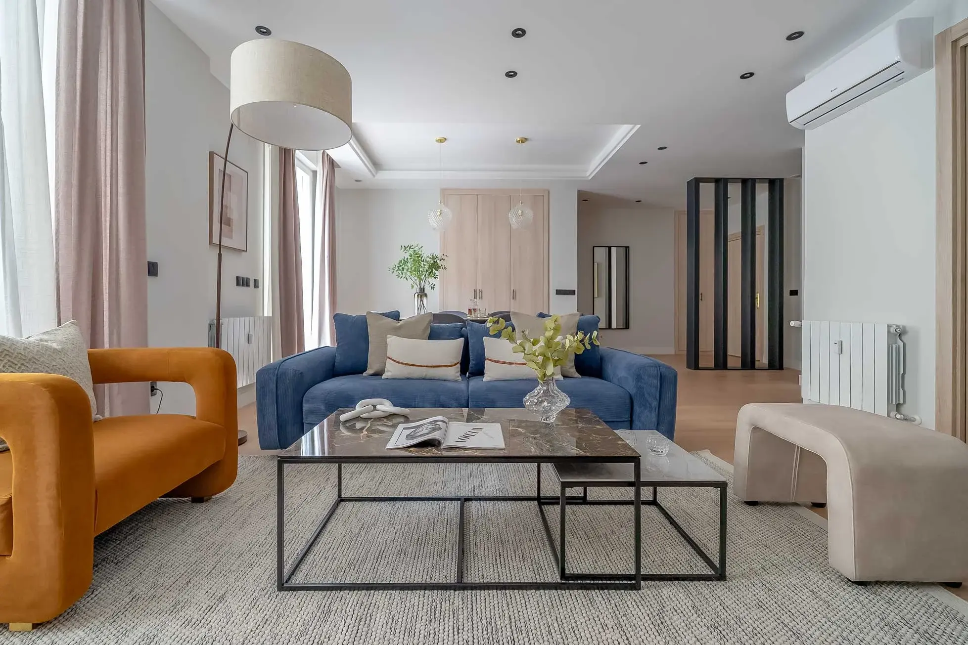Salón abierto y luminoso con un sillón curvo naranja, un sofá azul de pana con cojines, dos mesas nido de mármol oscuro y una lámpara de arco de gran pantalla.