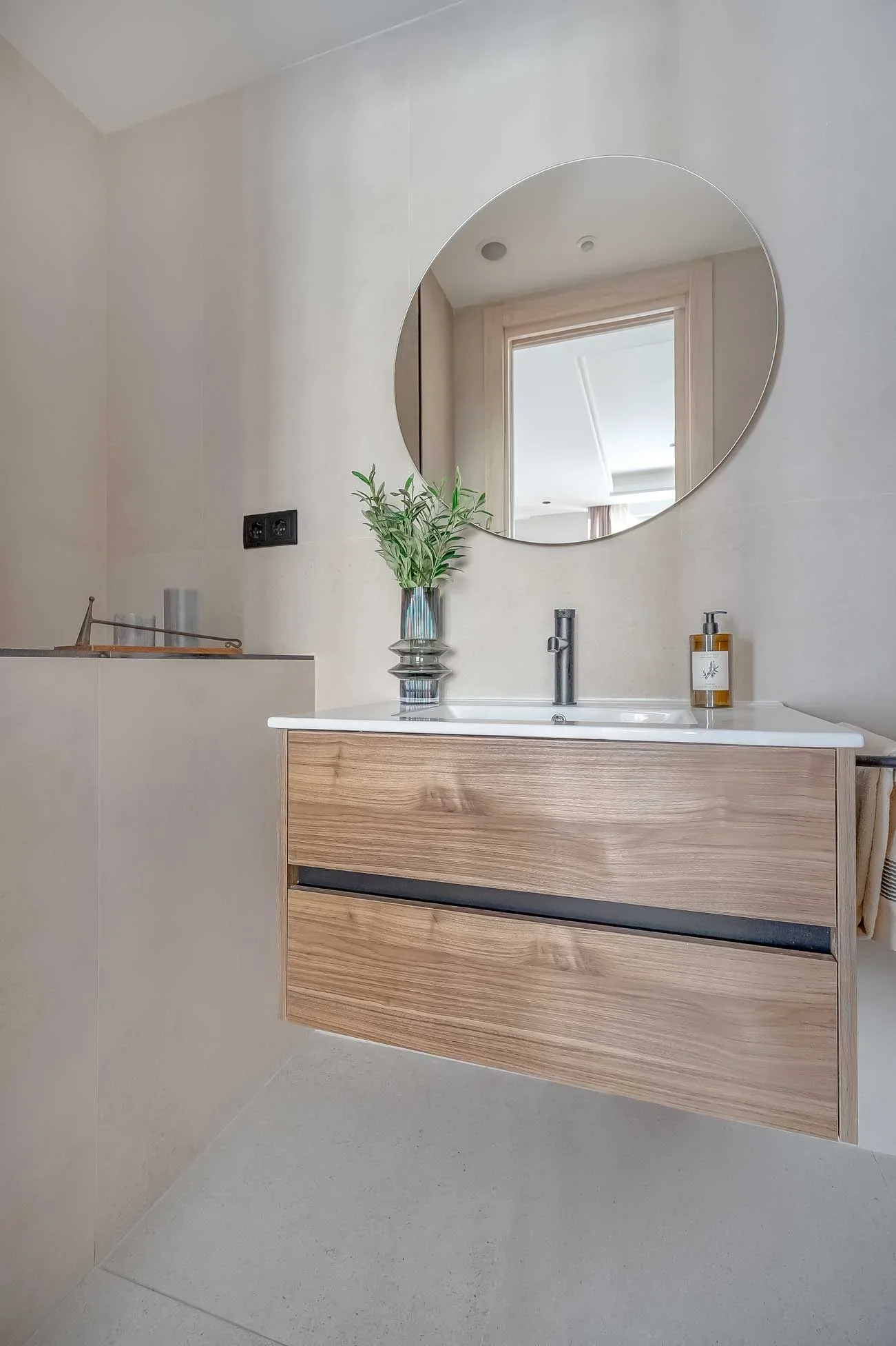 Aseo de cortesía con gran espejo redondo, mueble de lavabo de madera suspendido, grifería negra y una planta sobre la encimera.