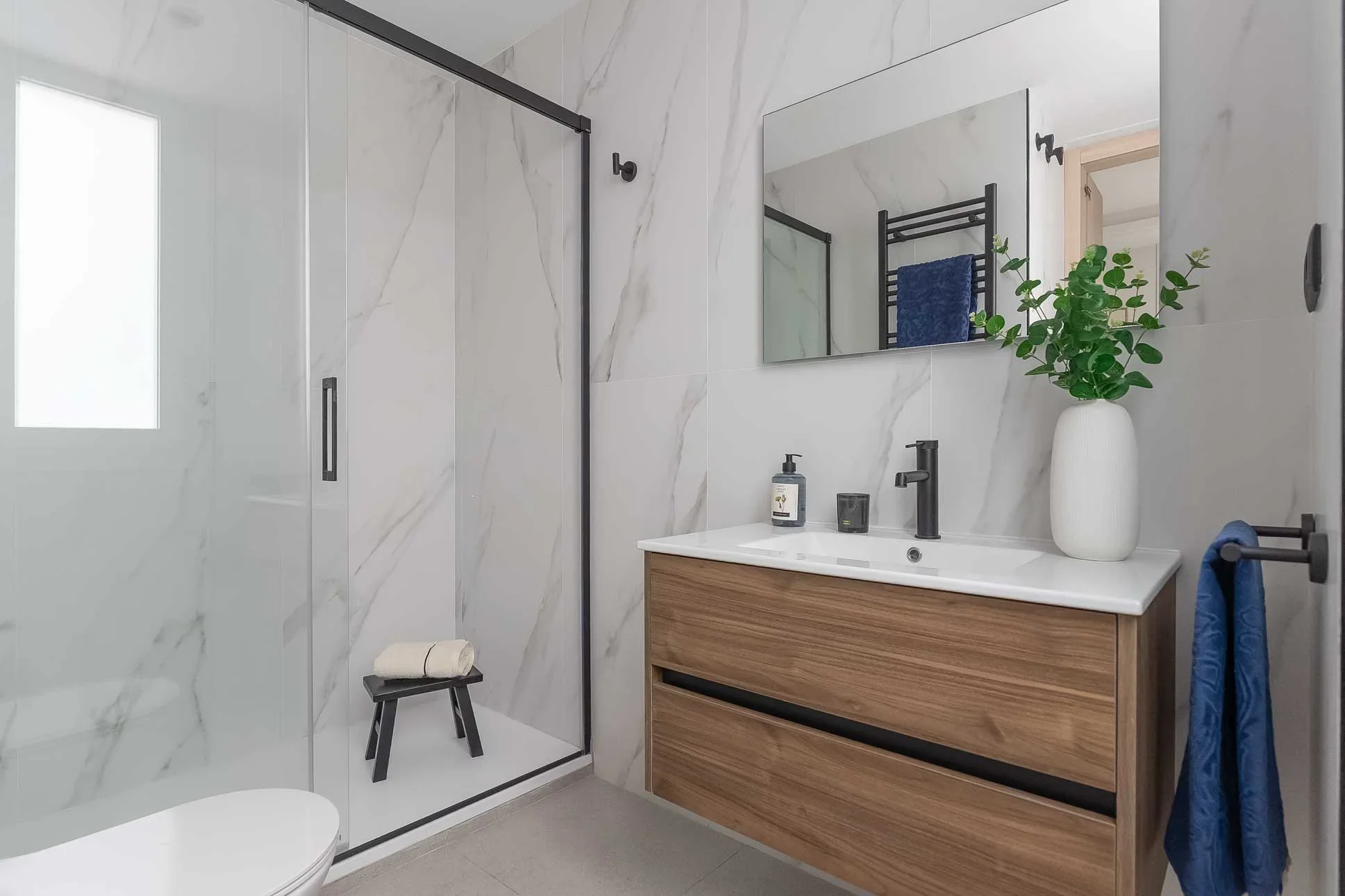 Baño revestido en mármol blanco con ducha de mampara de perfilería negra, mueble de madera con lavabo blanco y toalla azul en el toallero.