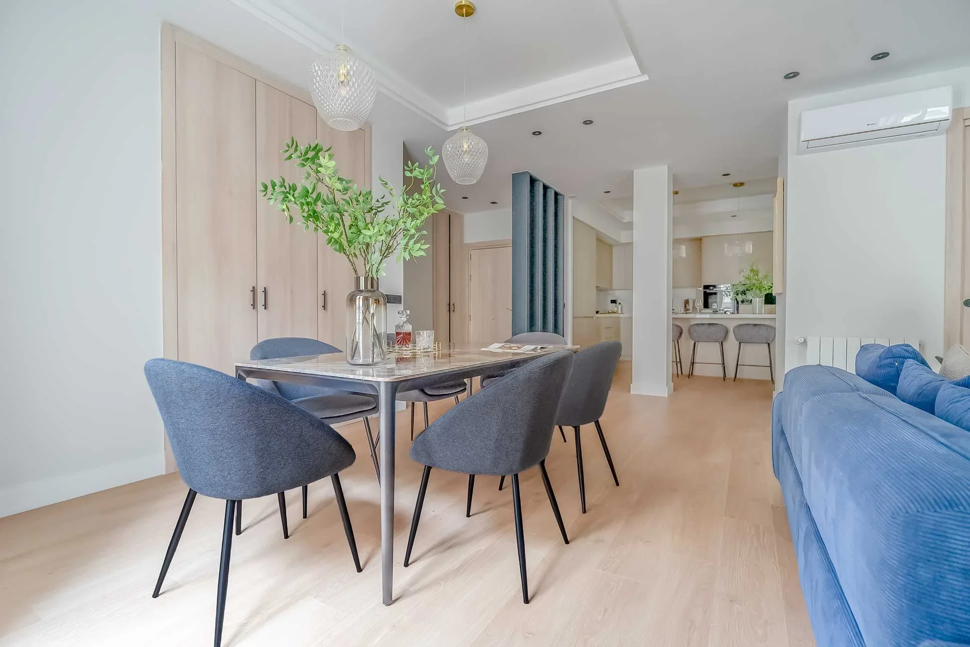 Comedor con mesa de mármol y patas metálicas, sillas tapizadas en gris azulado y conexión visual al fondo con la cocina.