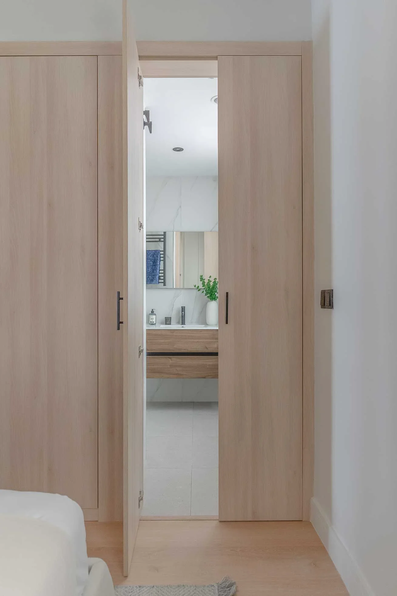 Puertas de armario de madera clara abiertas que conectan el dormitorio principal con su baño en suite revestido en mármol.