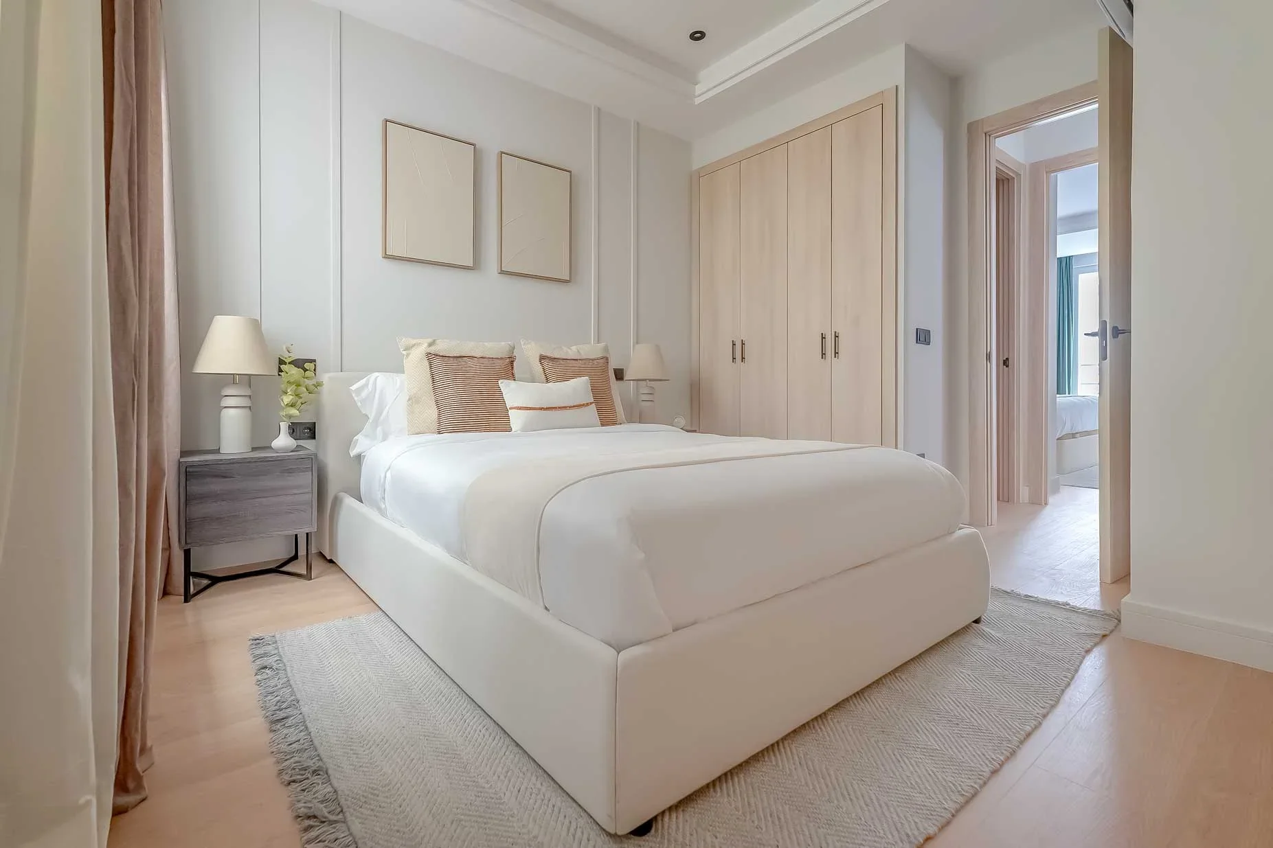 Dormitorio minimalista en tonos beige con cama vestida en blanco, cojines terracota, dos láminas abstractas y armarios de madera clara.