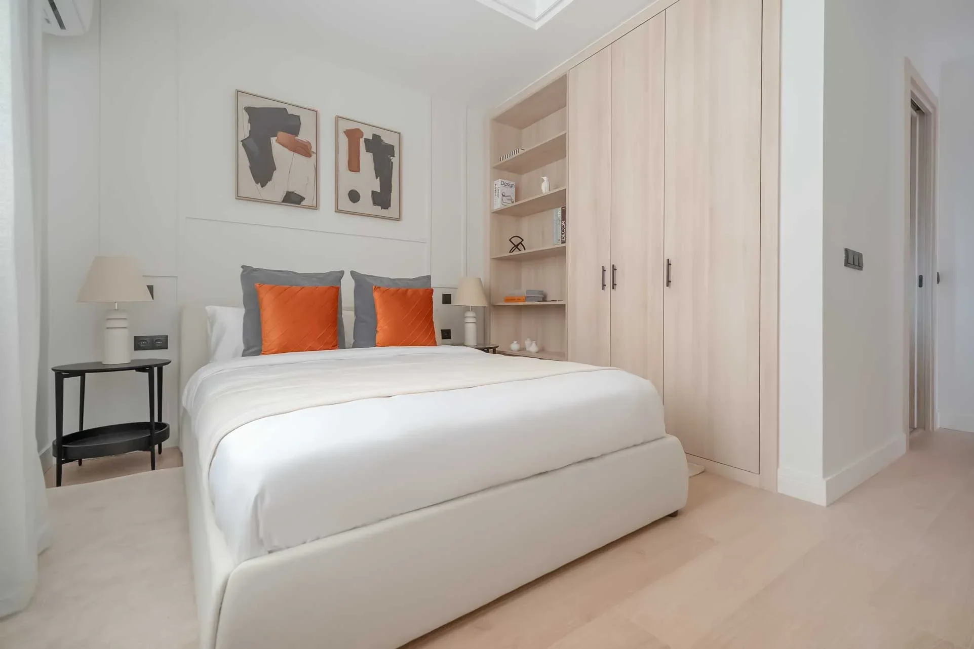 Dormitorio secundario con cama blanca, cojines grises y naranjas, láminas abstractas y estantería de madera integrada con libros y objetos de diseño.