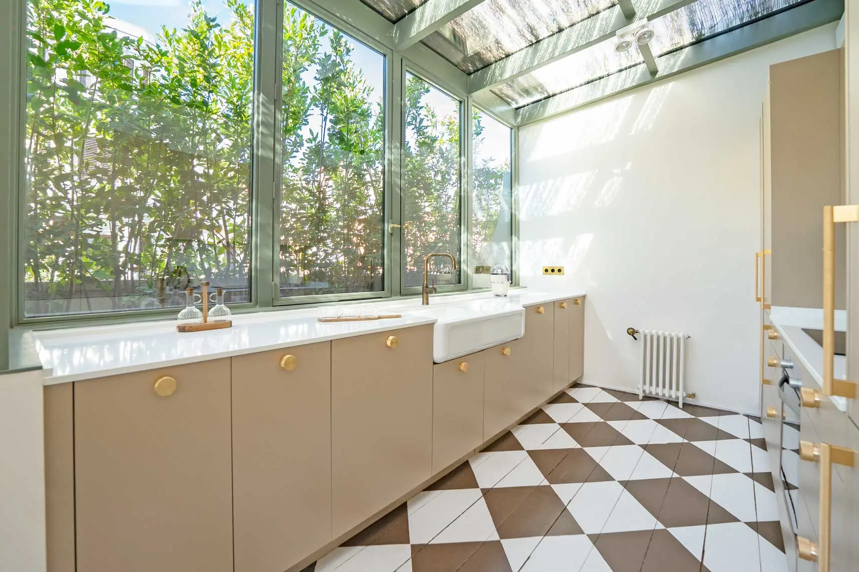 Cocina instalada en una galería acristalada de estructura verde, con muebles color arena, tiradores de latón, fregadero blanco y suelo damero en tonos marrón y blanco.