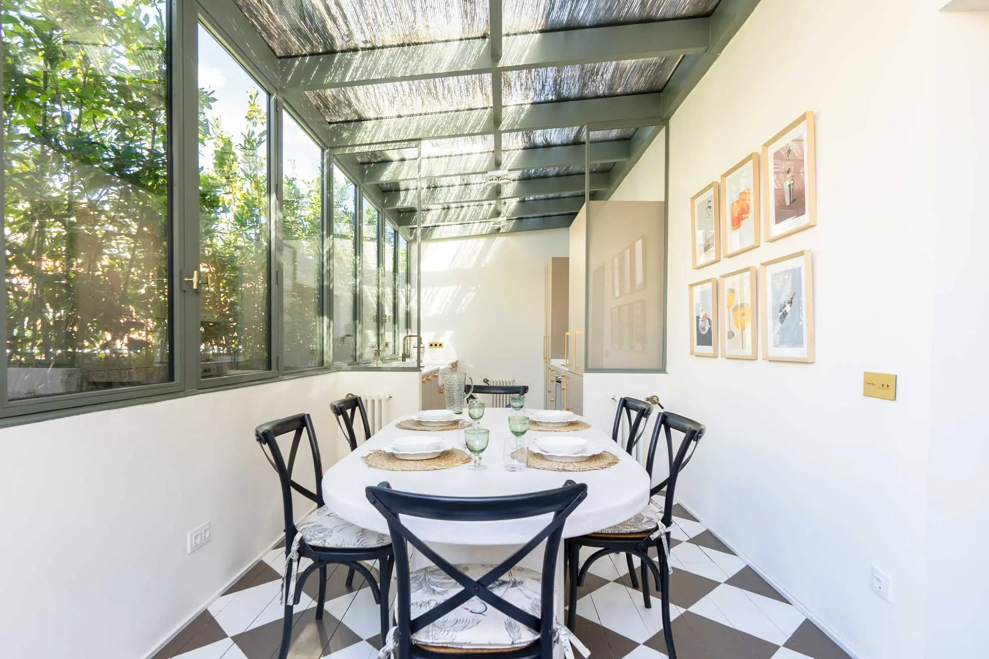 Comedor bajo la galería acristalada con mesa ovalada vestida, sillas negras de respaldo cruzado, una pared de láminas enmarcadas y suelo damero.