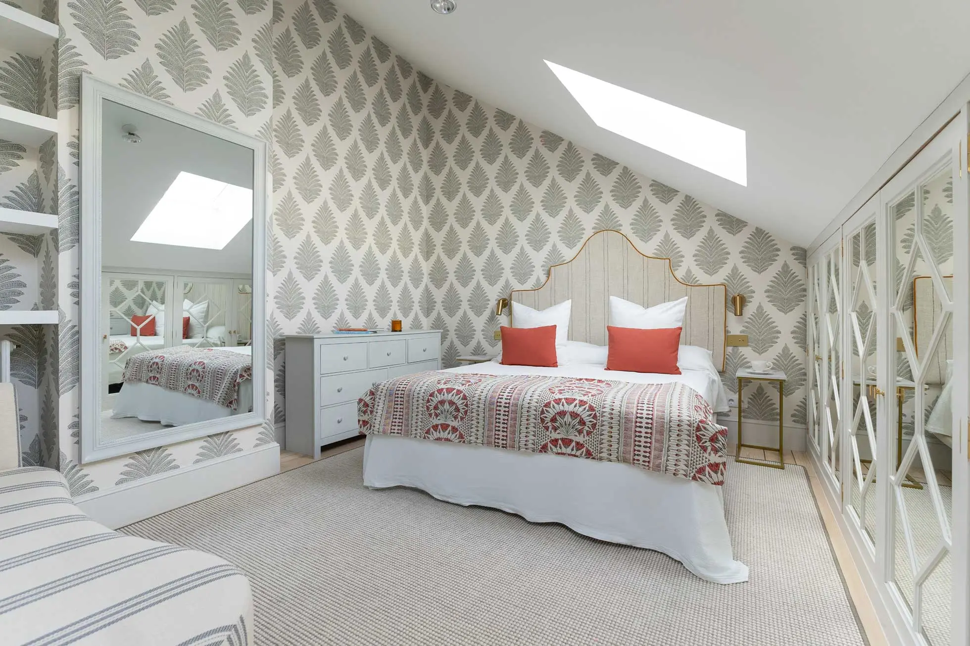 Dormitorio principal abuhardillado con papel pintado de hojas grises, cabecero tapizado con ribete dorado, cojines coral, tragaluz velux y armarios con espejo.