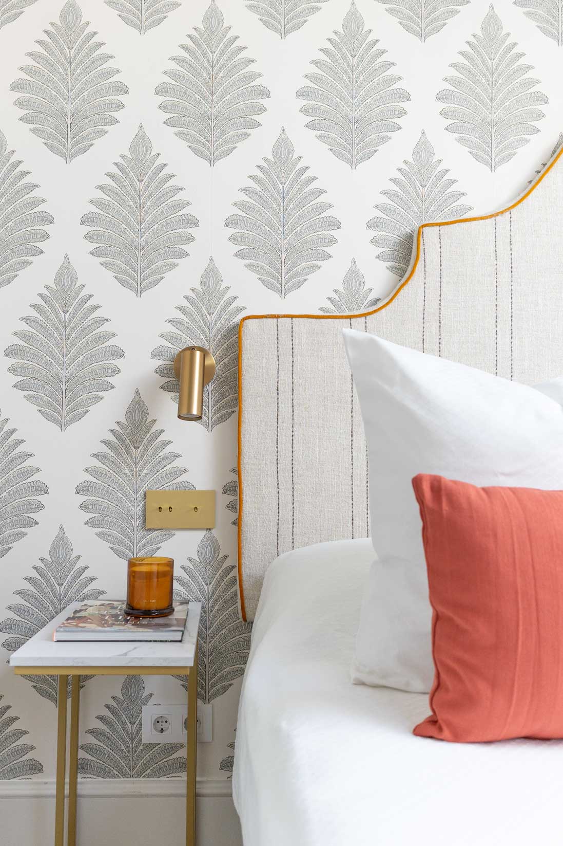 Detalle del dormitorio principal con papel pintado de palmeras, cabecero de lino a rayas con ribete mostaza, aplique de latón y mesita auxiliar de mármol.