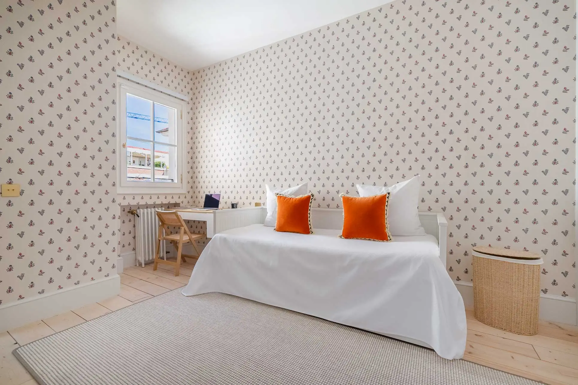 Segundo dormitorio con papel pintado de flores pequeñas, cama vestida en blanco con cojines naranjas, rincón de escritorio junto a la ventana y cesto de fibra natural.
