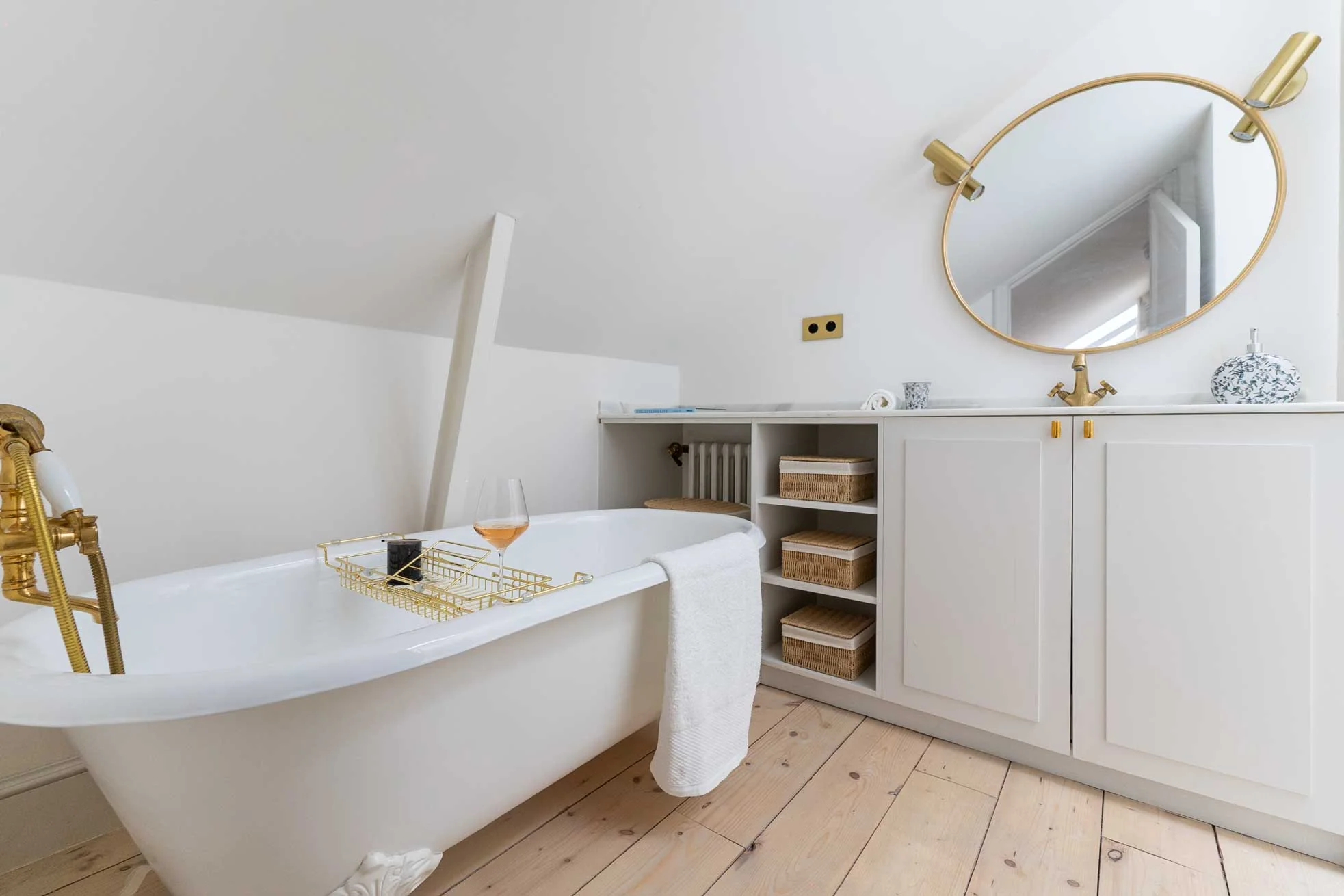 Baño abuhardillado con bañera exenta de estilo clásico, grifería dorada tipo teléfono, espejo redondo de latón y mueble bajo con cestas de mimbre.