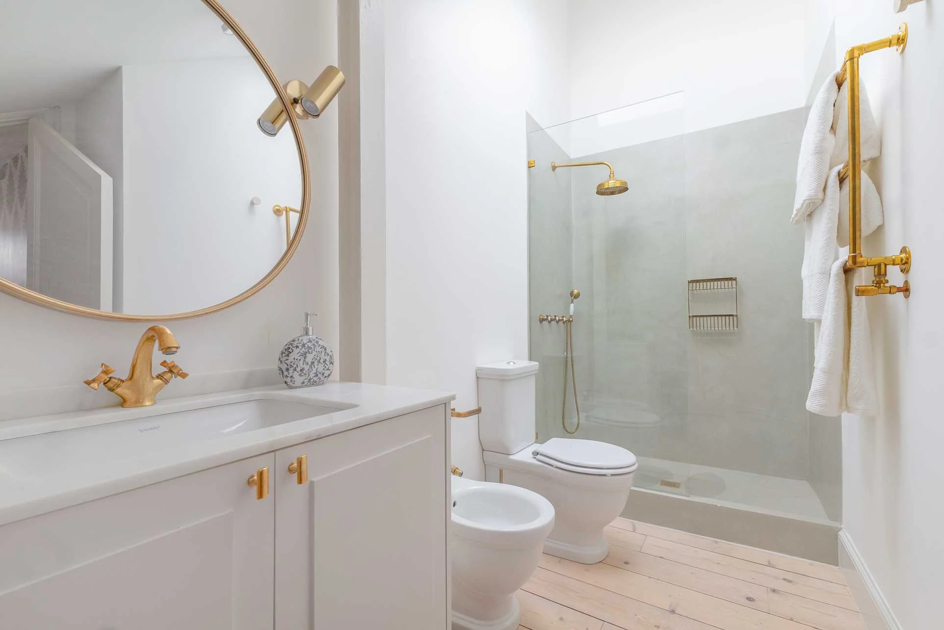 Baño con espejo redondo dorado, mueble blanco con grifería de latón, sanitarios clásicos y ducha de paredes de microcemento verde salvia con rociador de latón.