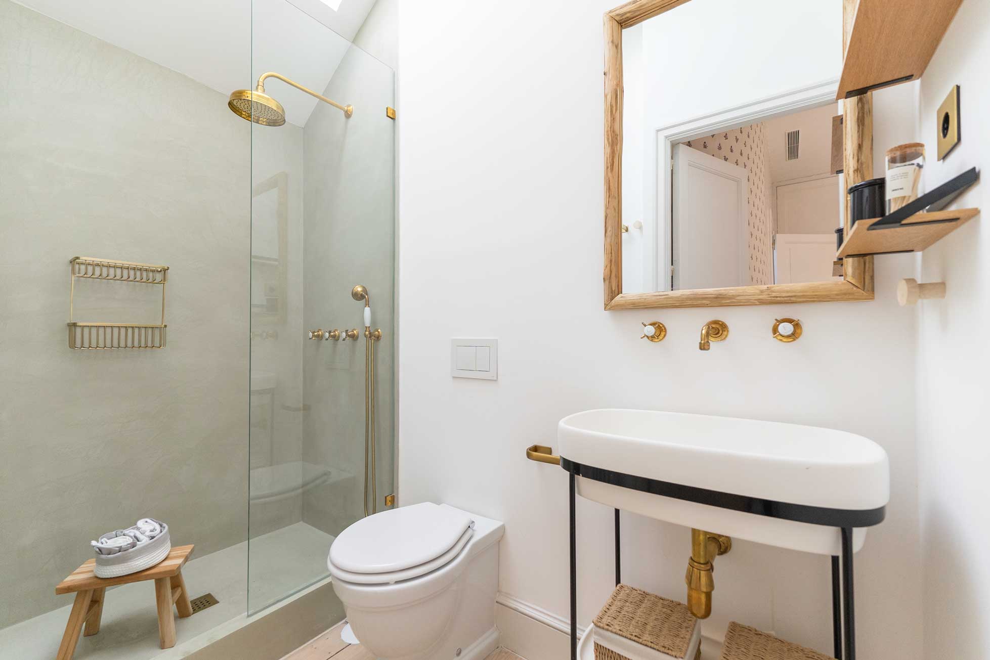 Aseo con ducha de microcemento verde salvia y mampara de cristal, rociador de latón, lavabo ovalado sobre consola metálica negra y espejo de madera.