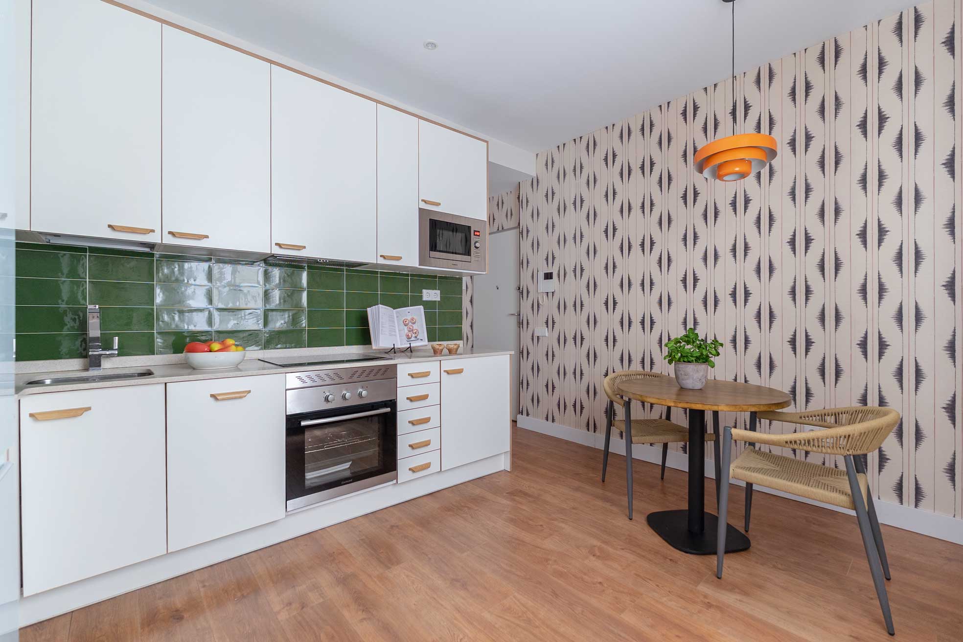 Cocina blanca con frente de azulejo verde botella brillante y rincón de office con mesa redonda de madera, sillas de fibra y papel pintado de motivo étnico en gris.