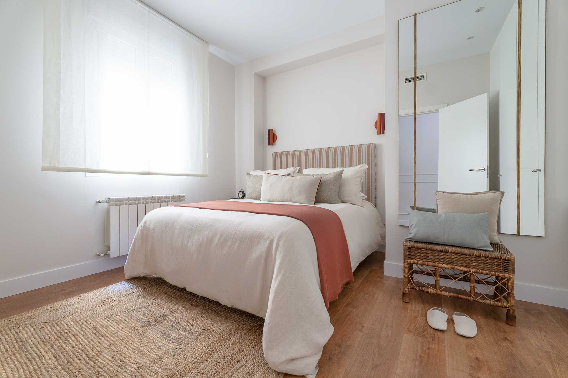 Dormitorio cálido con cabecero tapizado de rayas en tonos teja, pie de cama coral, alfombra de yute y espejo de pie sobre una banqueta de mimbre.