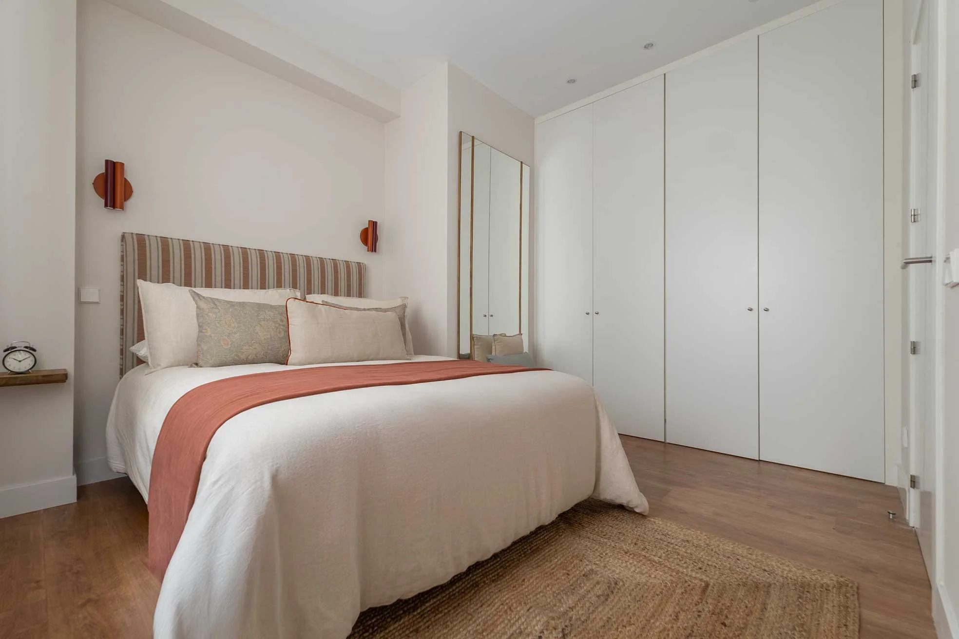 Otra vista del dormitorio con el cabecero de rayas, armarios empotrados blancos, apliques de pared en color teja y alfombra de yute.