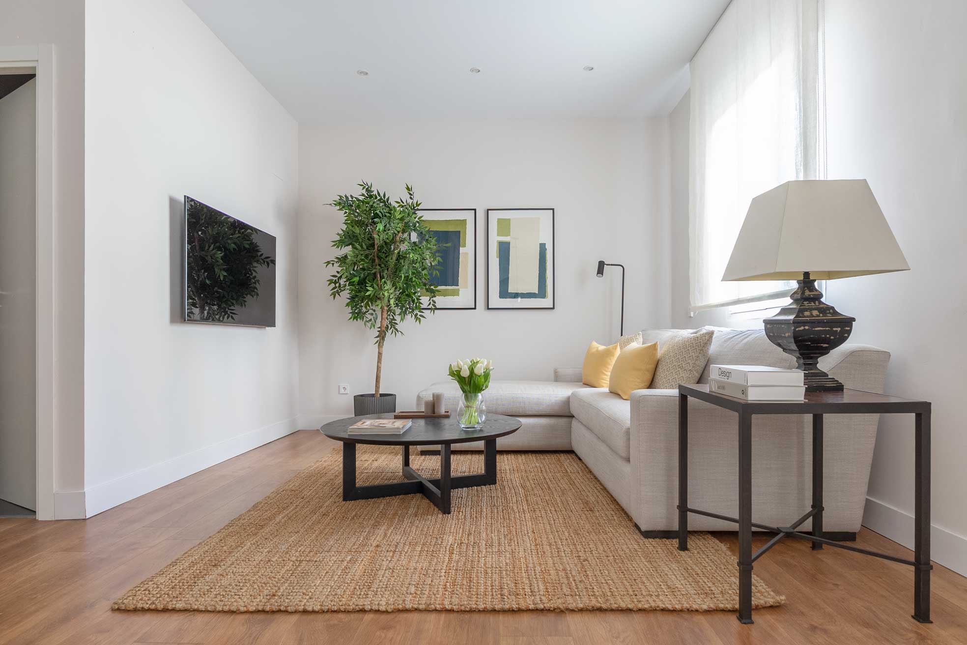 Otra perspectiva del salón hacia la televisión integrada en la pared, con la planta, las láminas abstractas y la mesa de centro redonda oscura.