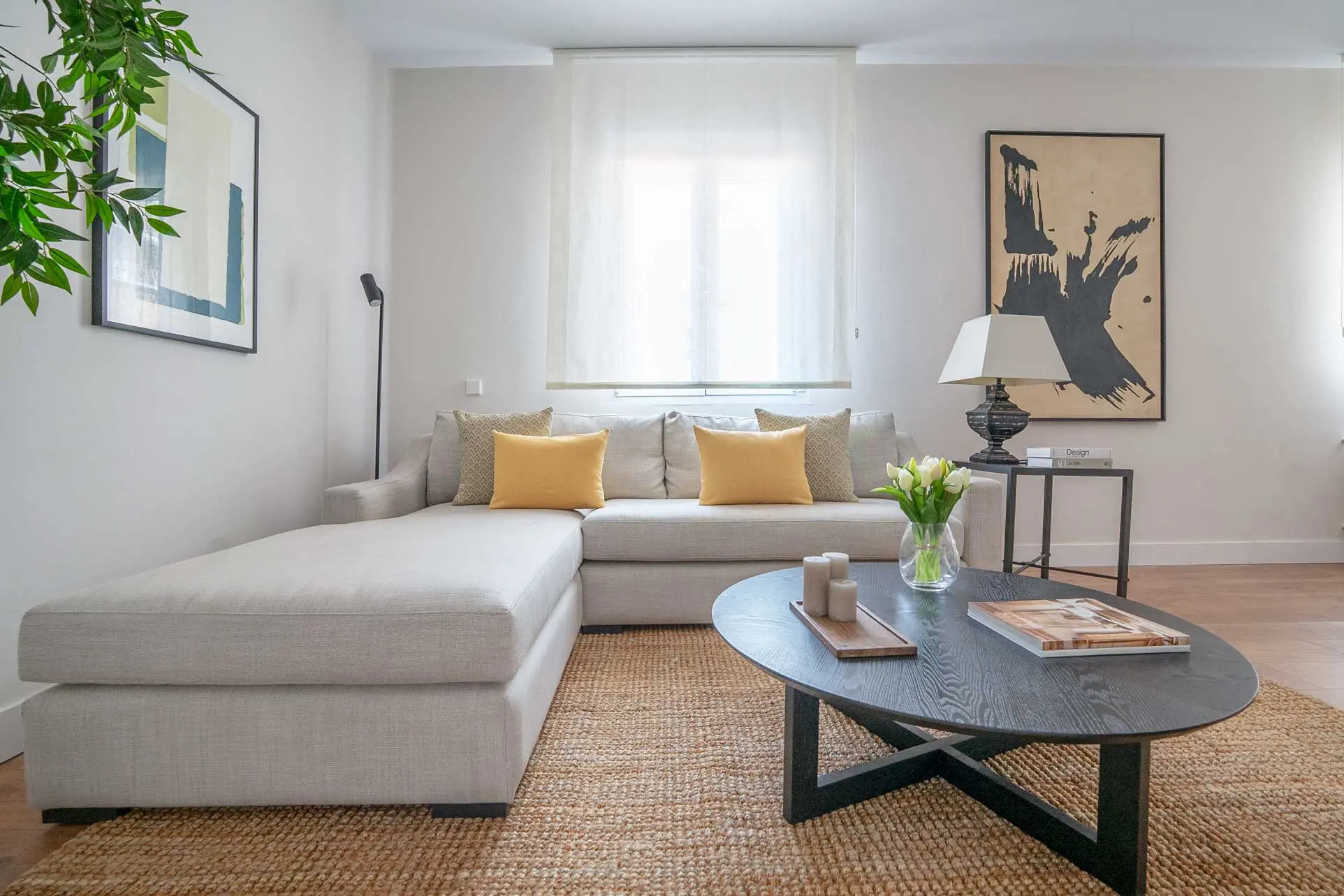 Plano del sofá chaise longue beige con cojines mostaza, mesa de centro ovalada negra y la obra abstracta en negro al fondo.