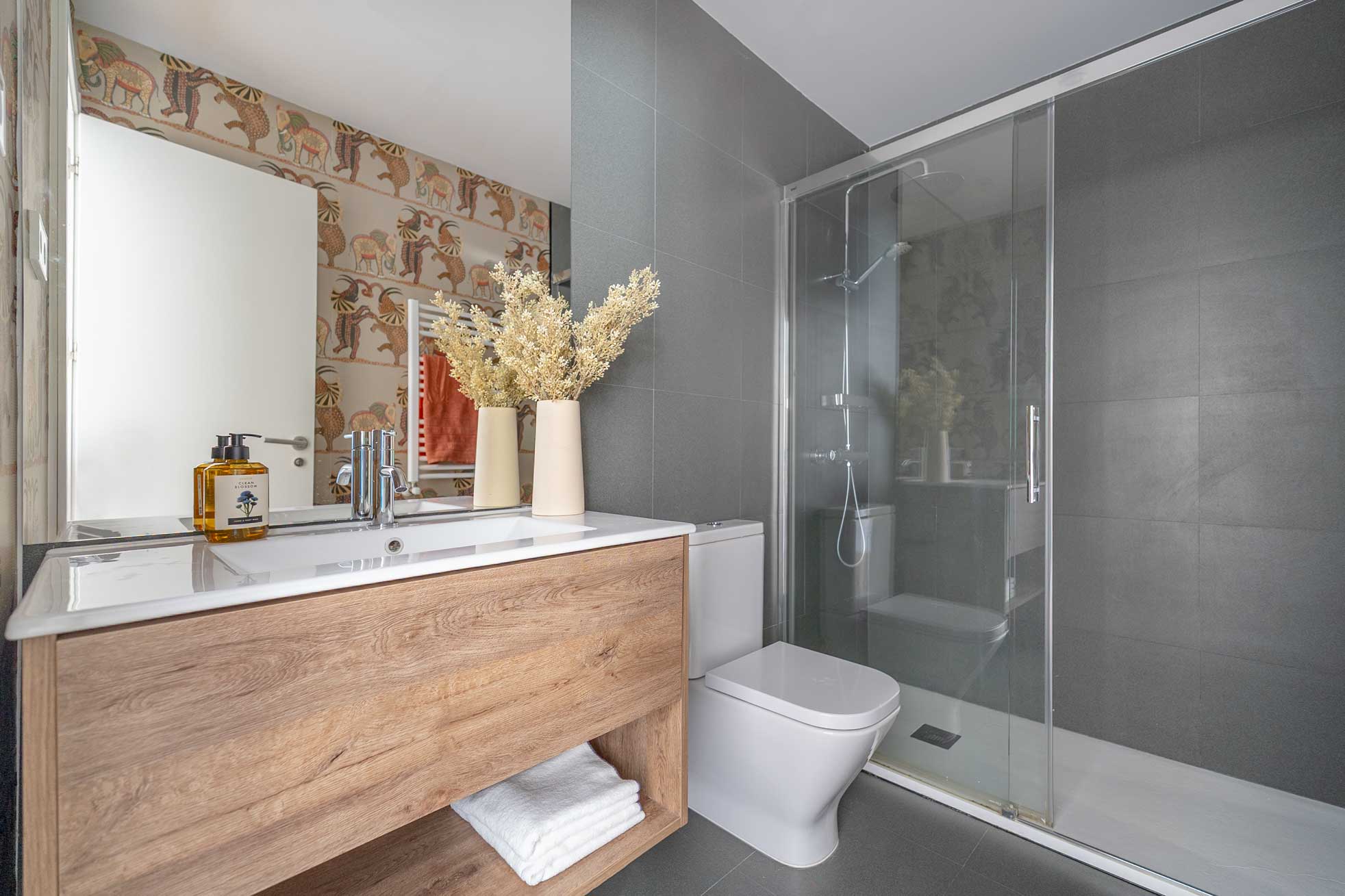 Baño con mueble de madera y lavabo blanco, papel pintado de elefantes, ducha de gres gris con mampara de cristal y toques en color teja.