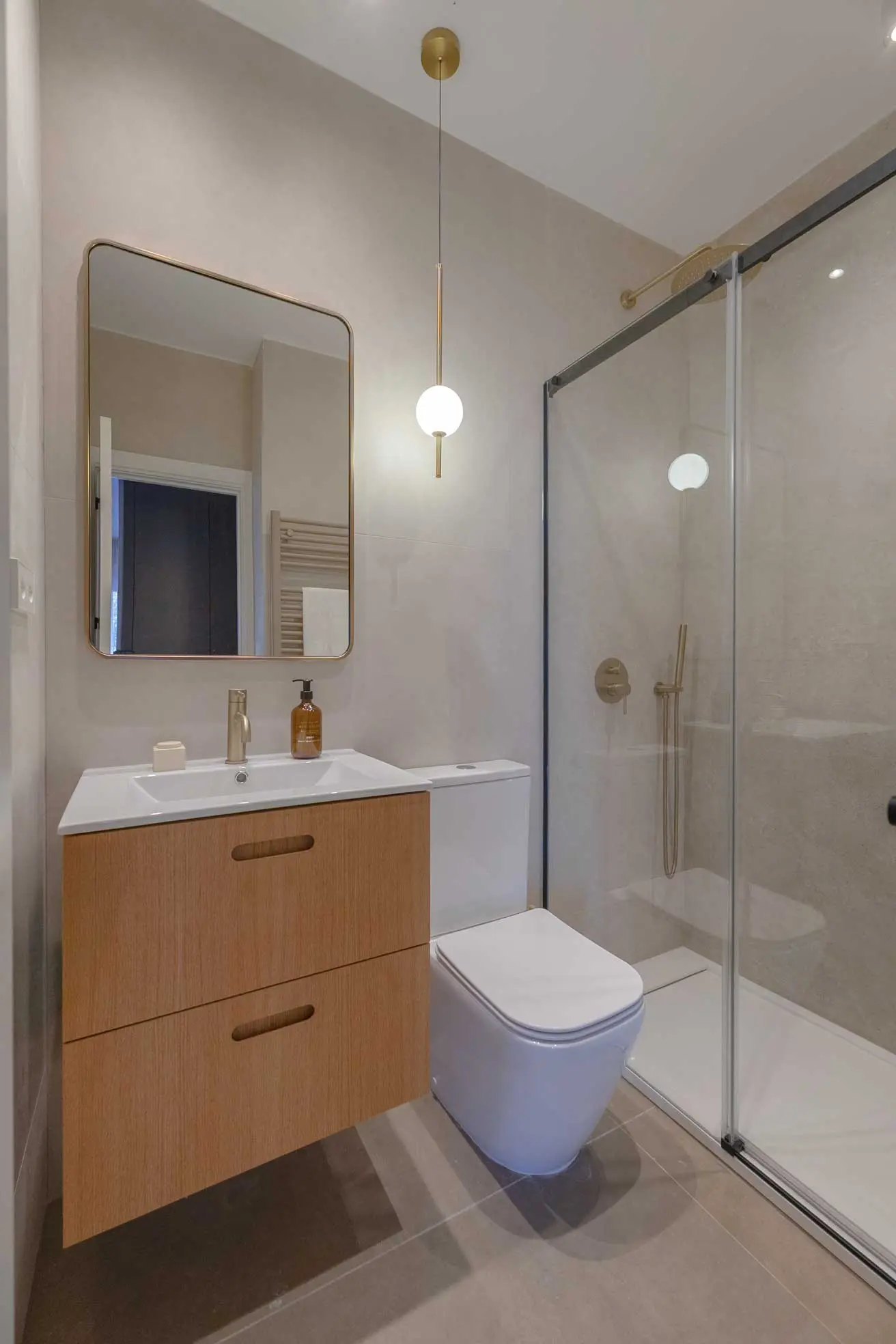 Baño con mueble de lavabo suspendido en madera clara, espejo de marco dorado, lámpara colgante esférica y ducha con mampara de cristal y grifería dorada sobre gres beige.