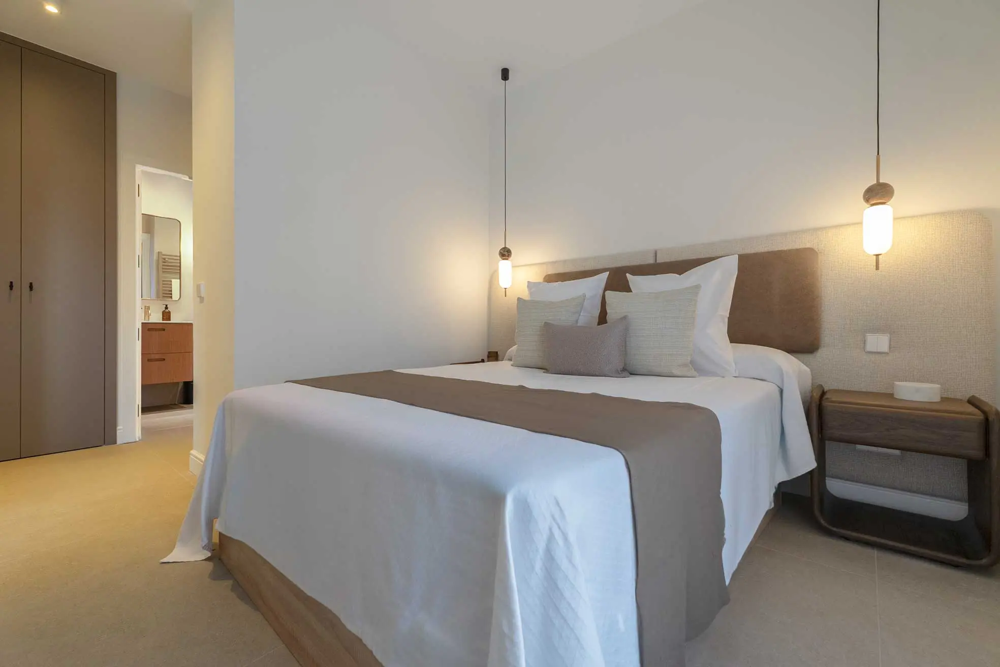 Dormitorio principal minimalista con cabecero tapizado de perfil bajo, ropa de cama en blanco y tonos tierra, dos lámparas colgantes esféricas y vistas al baño en suite.