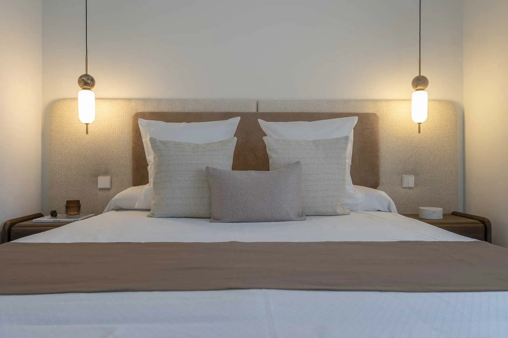 Plano frontal y simétrico de la cama de matrimonio con cabecero tapizado, mesillas de madera y dos lámparas colgantes de diseño a juego.