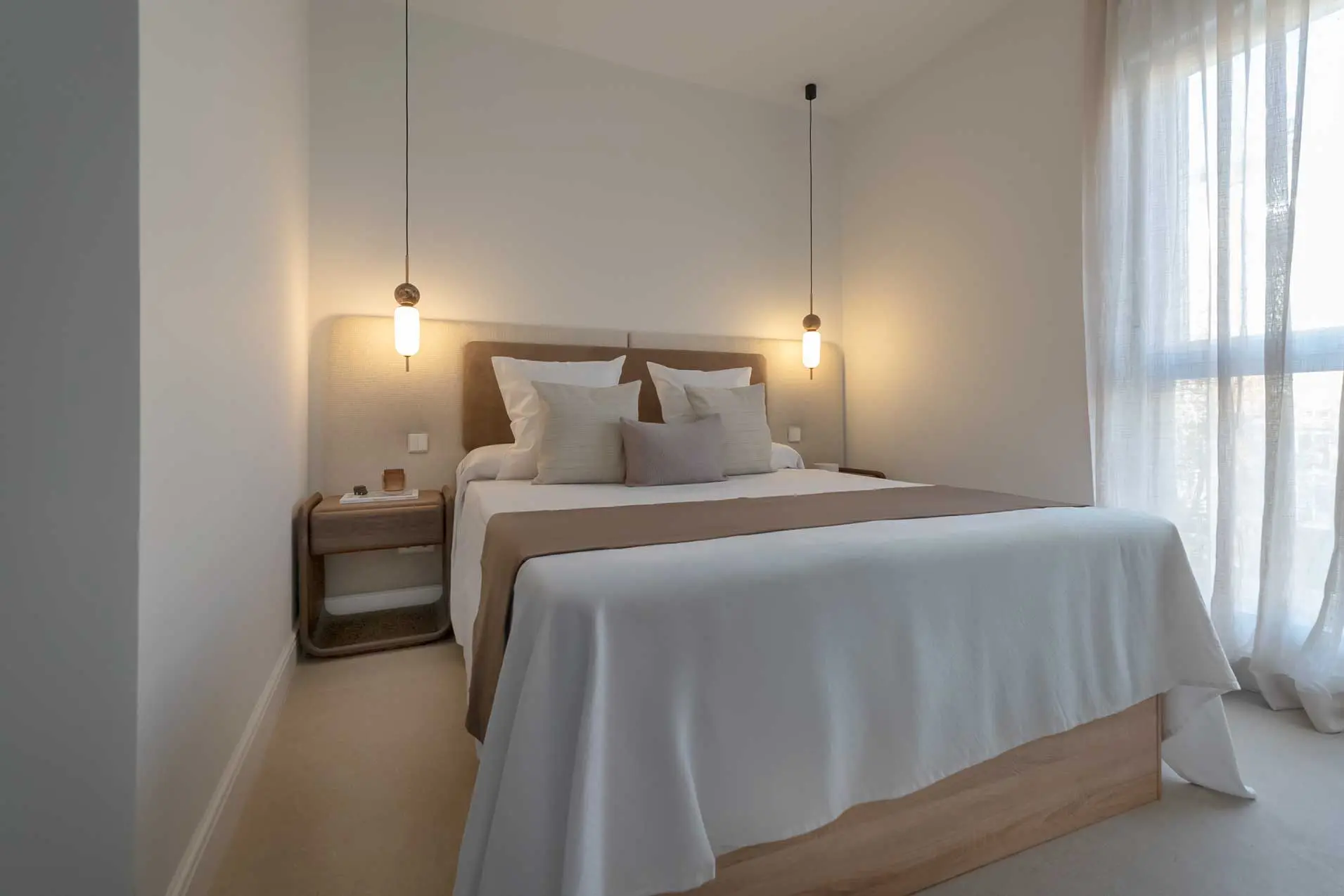 Perspectiva del dormitorio desde la ventana, con la cama vestida en tonos neutros, las lámparas colgantes y un gran ventanal con visillos que aporta luz natural.