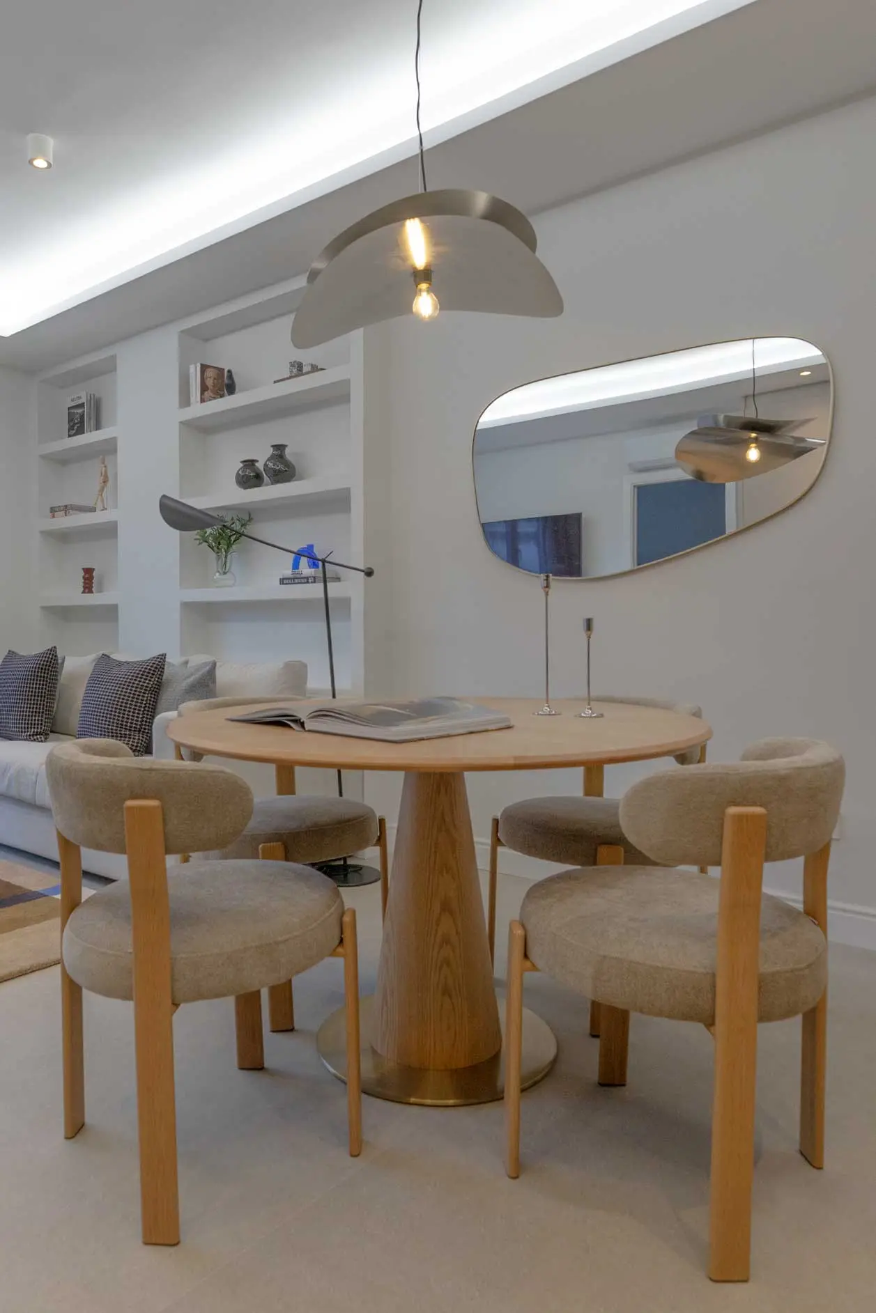 Zona de comedor con mesa redonda de madera y base dorada, sillas tapizadas en tonos arena, espejo orgánico asimétrico y lámpara de techo escultórica metálica.