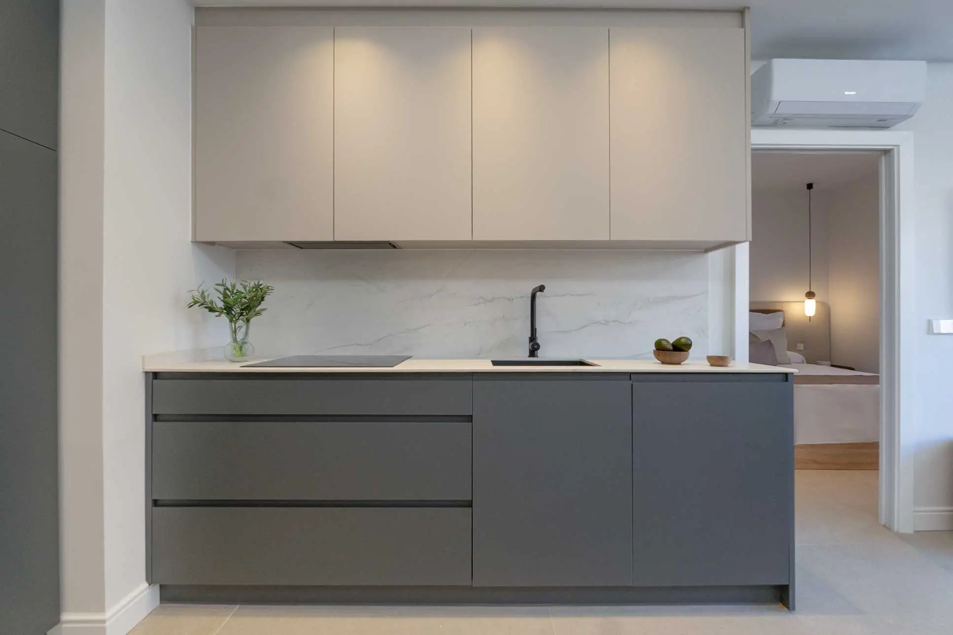 Cocina de diseño con módulos inferiores en gris antracita, muebles altos en beige, encimera y frente de mármol blanco y grifería negra, abierta al dormitorio.