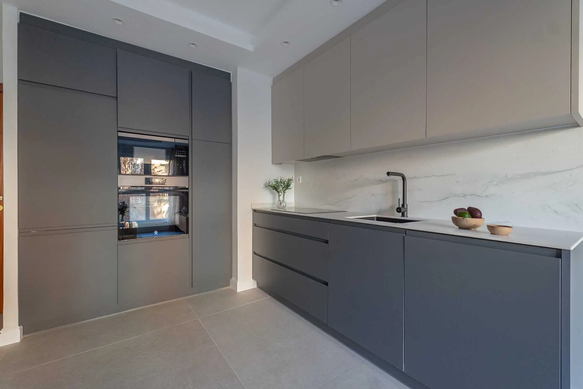 Perspectiva de la cocina con la columna de horno integrada en gris antracita, muebles altos beige, encimera de mármol blanco y suelo de gres gris.