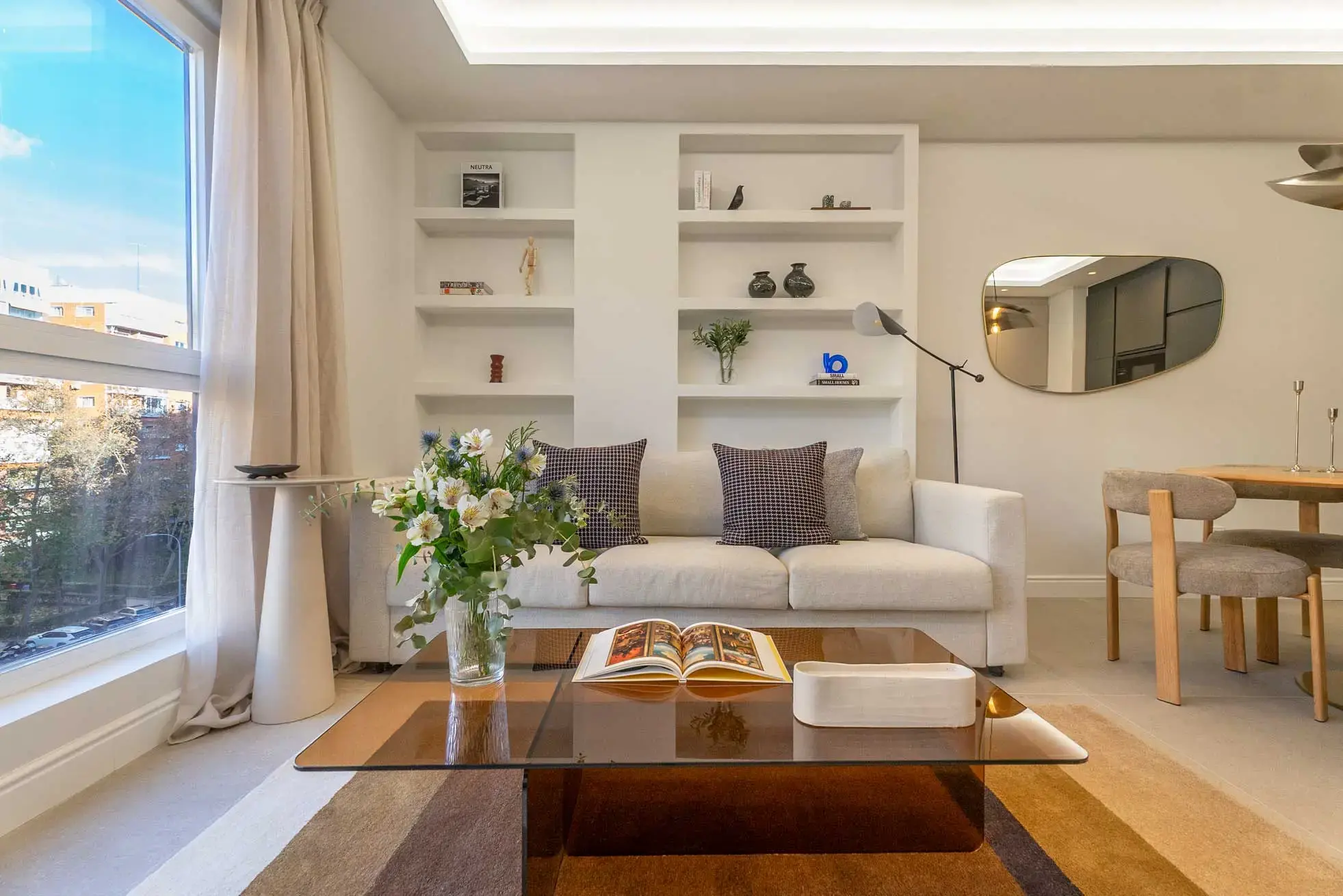 Salón luminoso con sofá beige de cojines de cuadros, mesa de centro baja de cristal ahumado, estantería de obra blanca decorada y espejo orgánico.