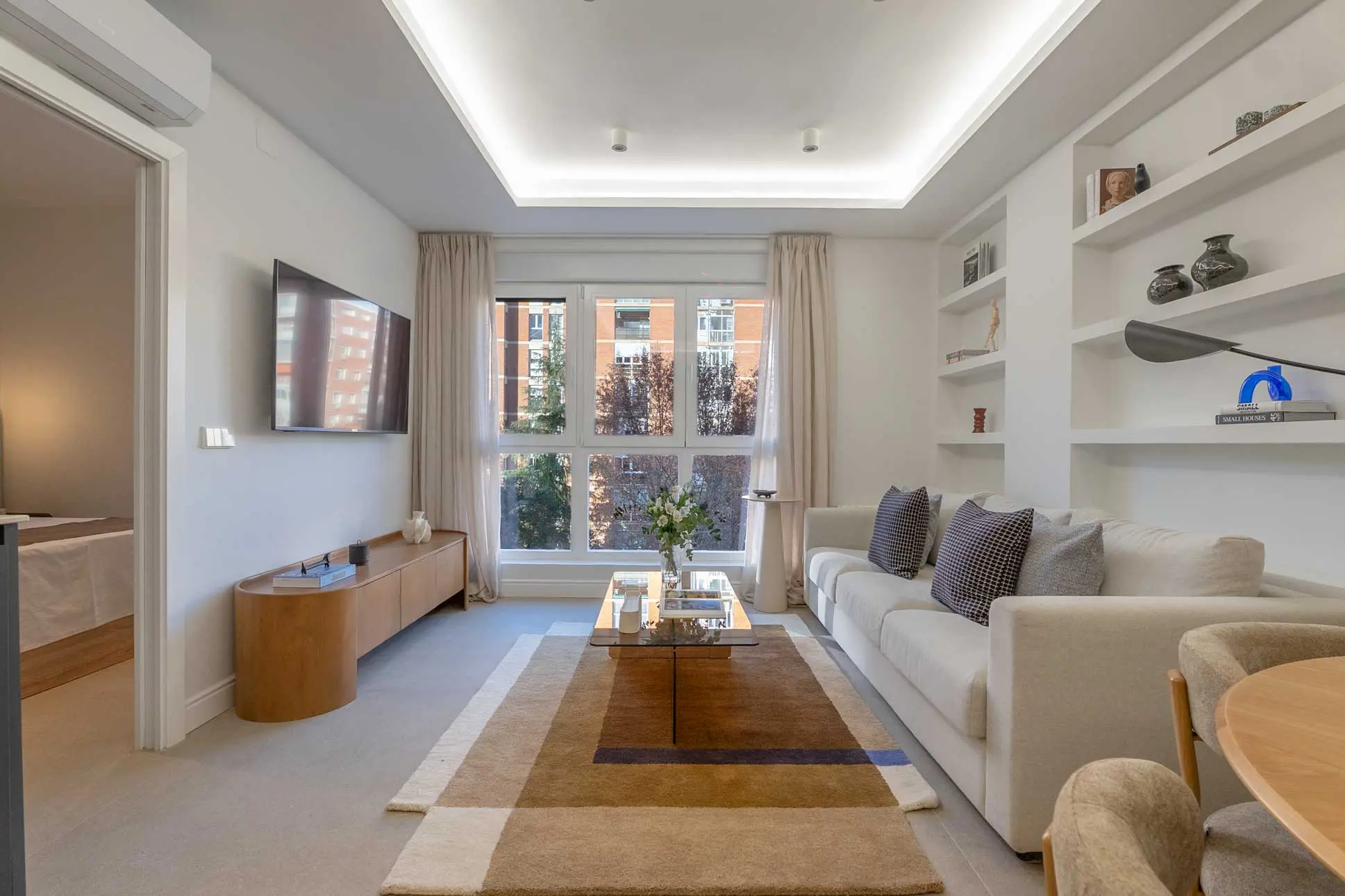 Perspectiva del salón hacia la televisión de pared y un mueble bajo de madera de líneas curvas, con salida al dormitorio.
