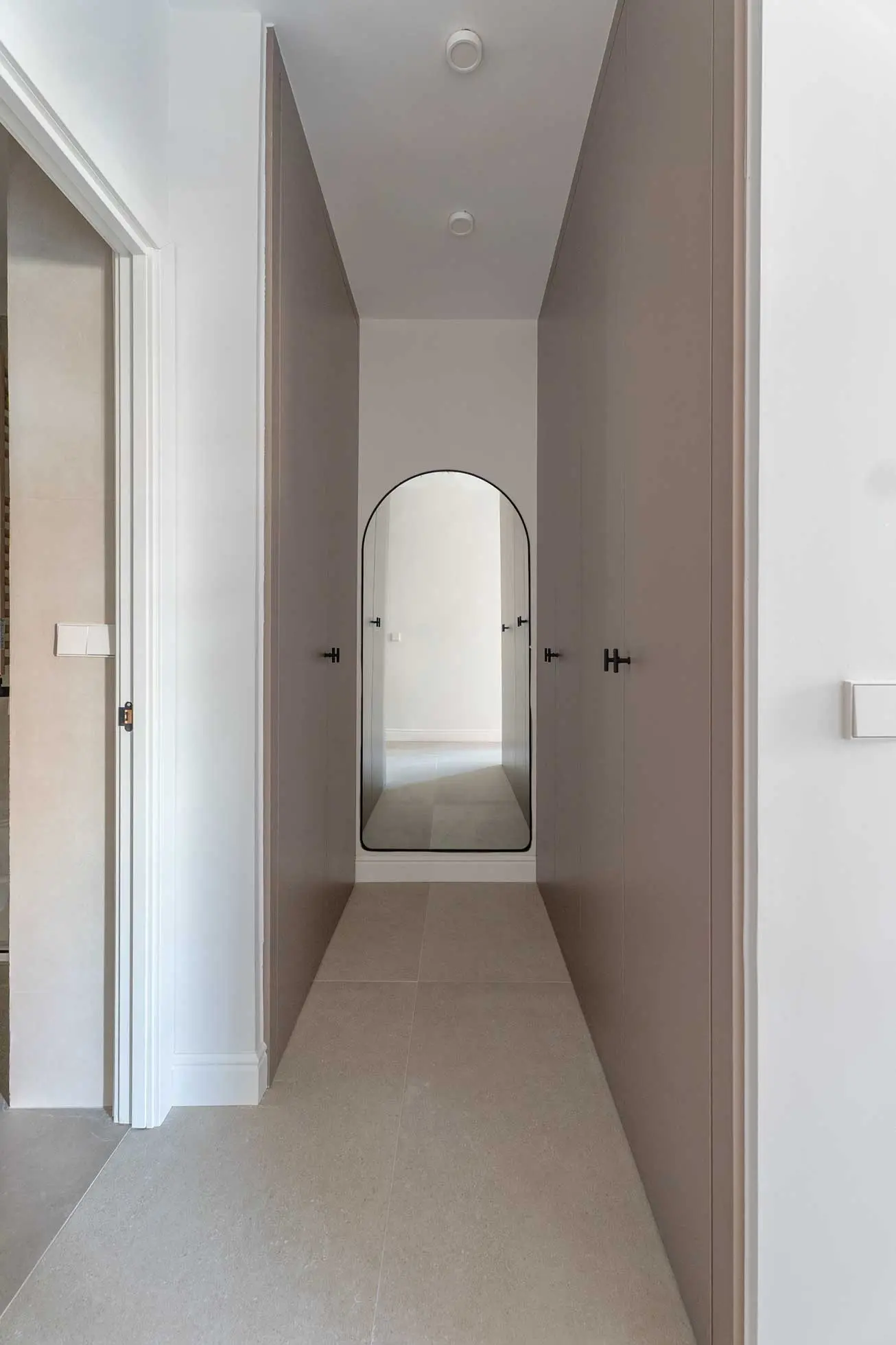 Pasillo distribuidor flanqueado por armarios empotrados lacados en color topo, con un espejo de cuerpo entero en forma de arco al fondo.