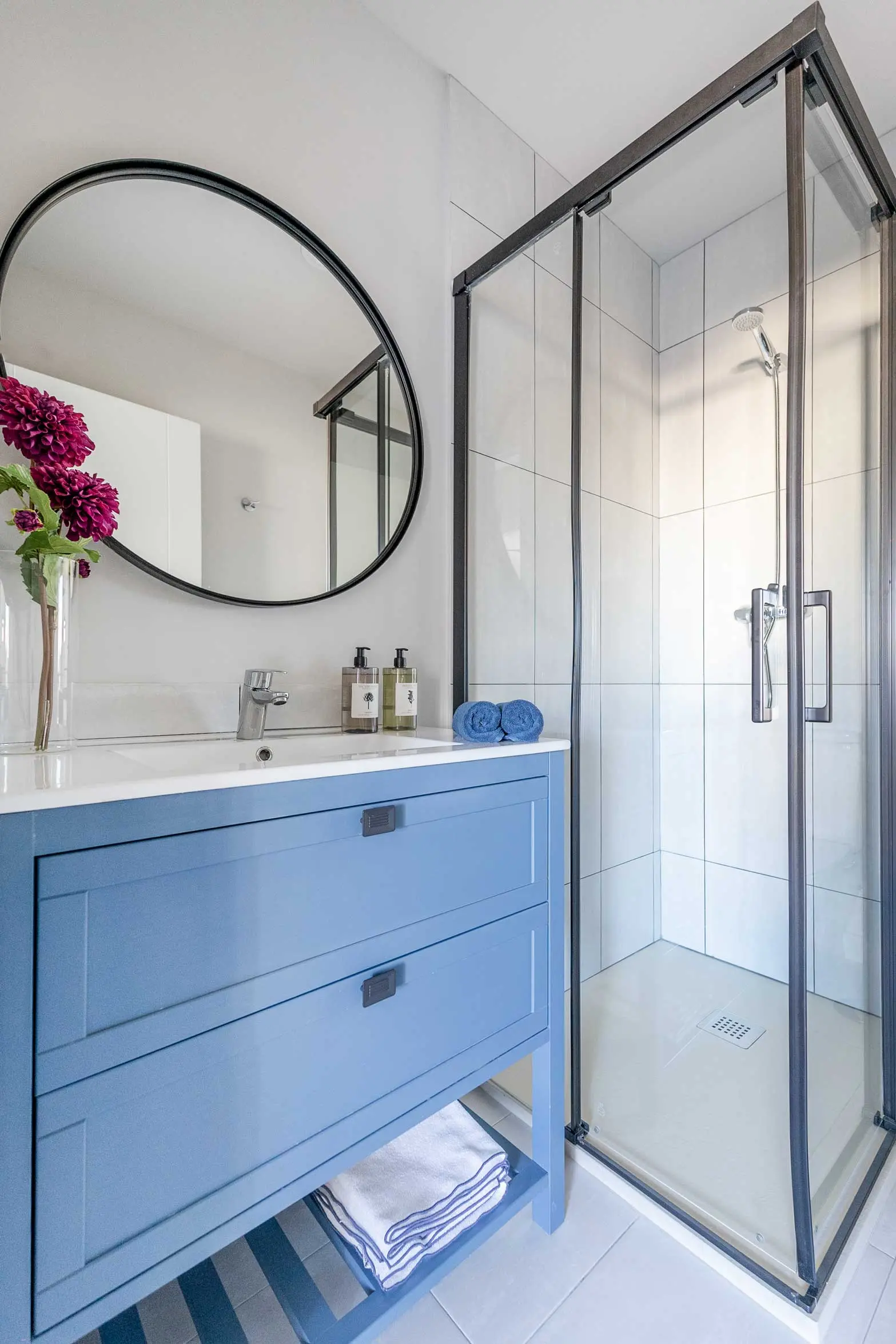 Primer cuarto de baño con mueble bajo lavabo en color azul pastel, espejo redondo y mampara con perfilería negra