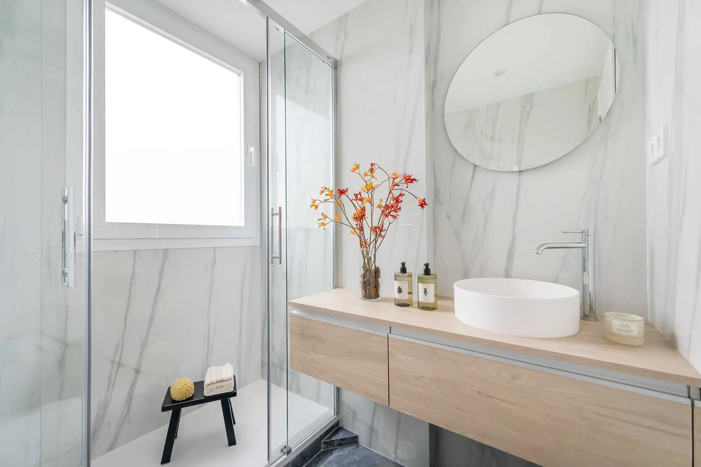 Cuarto de baño de diseño moderno con revestimiento de mármol blanco, lavabo sobre encimera de madera y espejo redondo