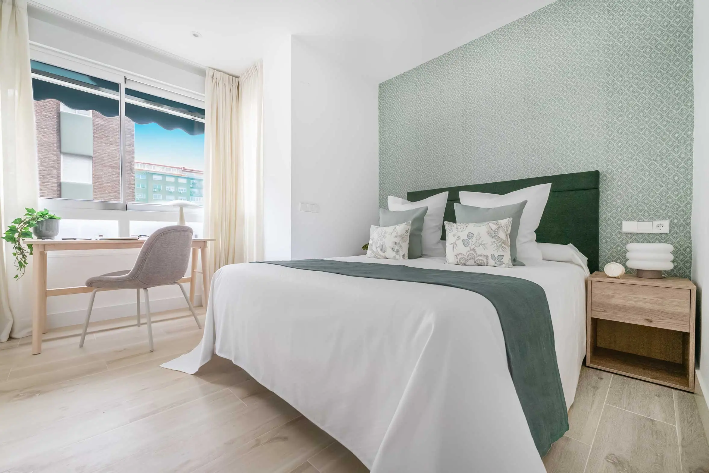 Dormitorio principal con una cama vestida en tonos blancos y verdes, cabecero tapizado y rincón de escritorio al fondo