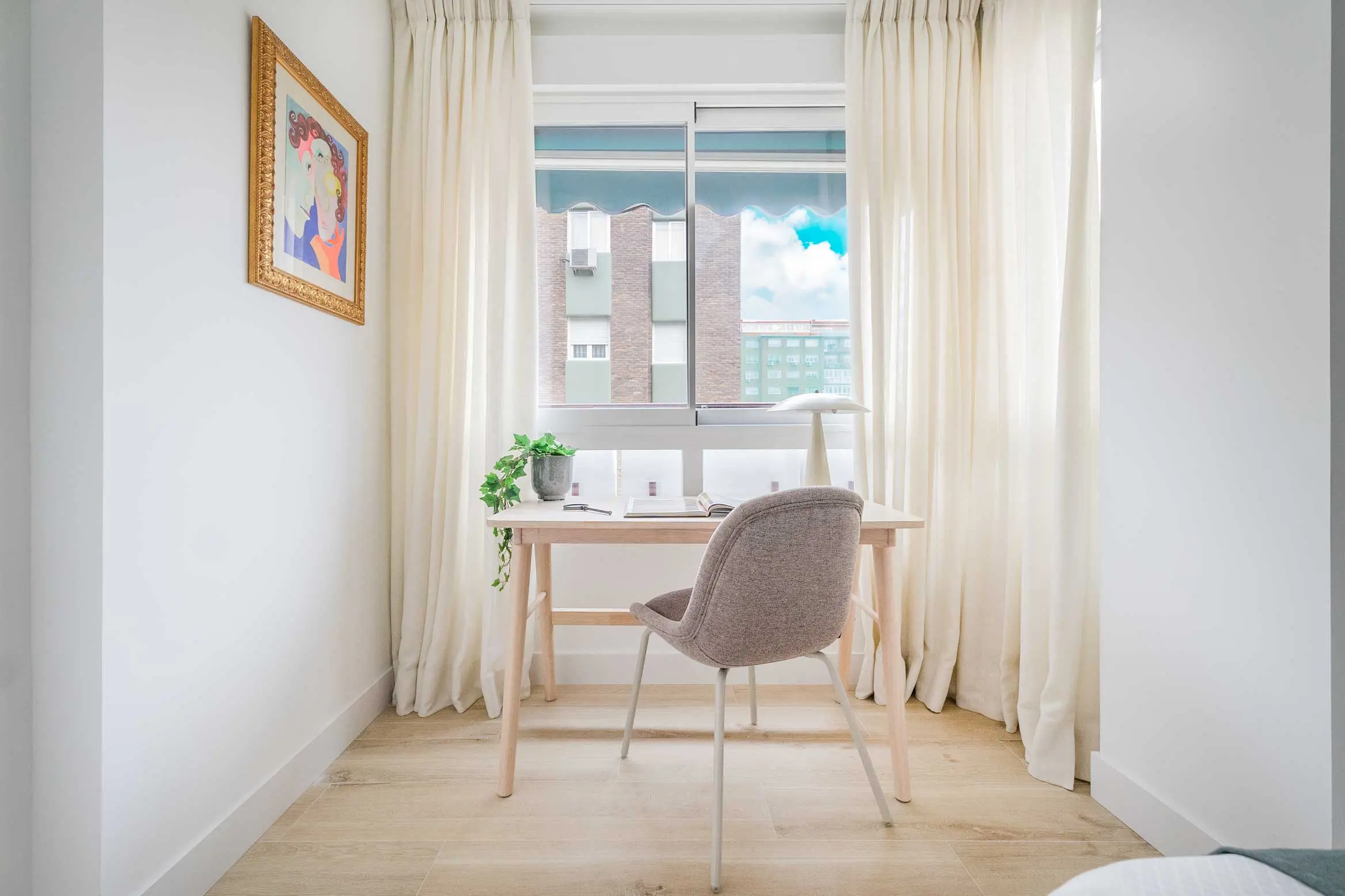 Detalle del rincón de trabajo junto a la ventana del dormitorio con mesa de madera clara y silla tapizada gris
