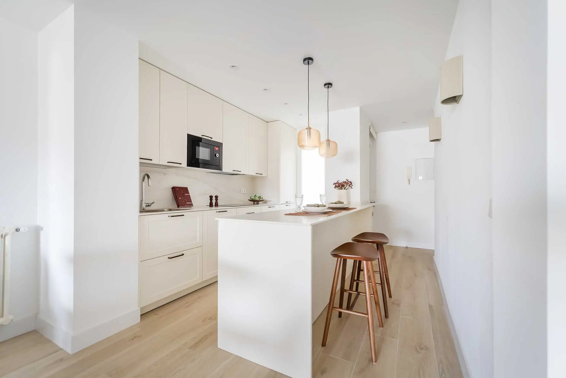 Perspectiva luminosa de la cocina minimalista blanca con isla central, taburetes de madera y lámparas de suspensión de diseño en el apartamento Boix y Morer