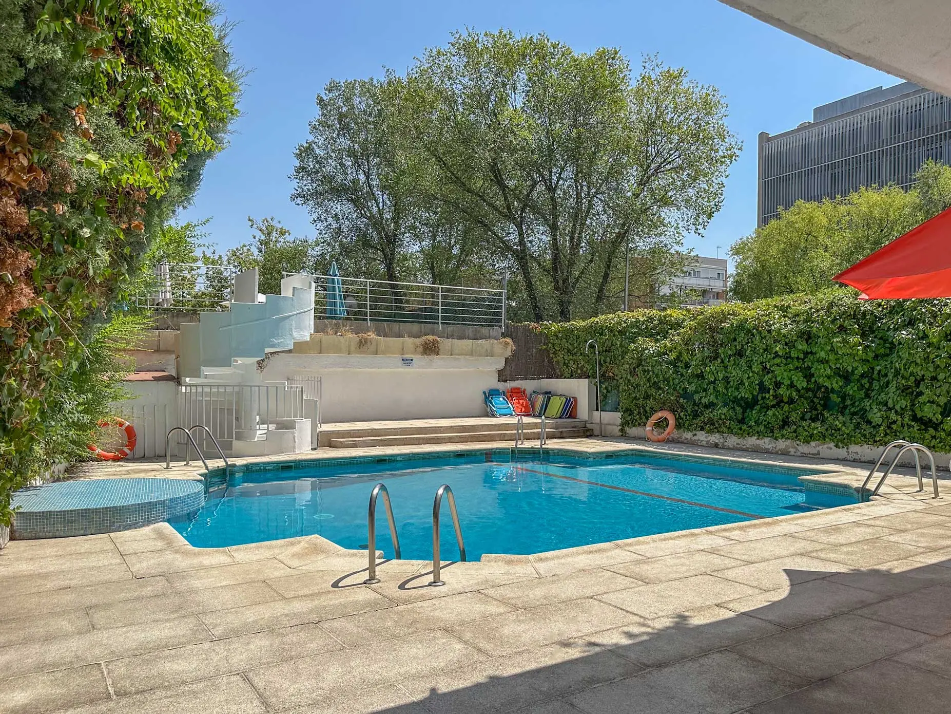 Ángulo abierto de la piscina comunitaria rodeada de vegetación frondosa y zonas de descanso pavimentadas