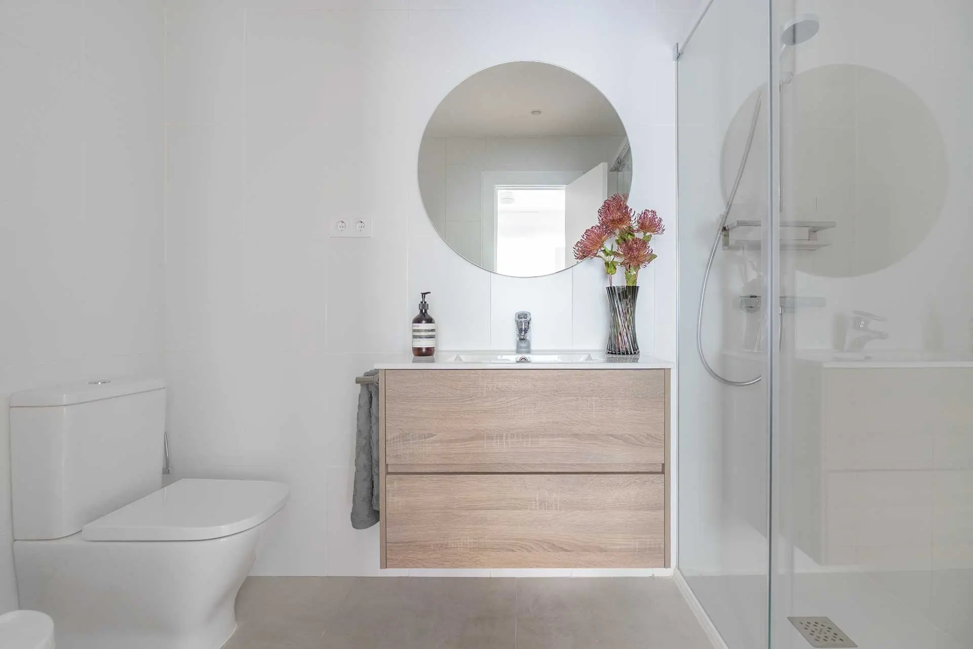 Cuarto de baño moderno con mueble suspendido de madera clara, lavabo integrado, espejo circular y mampara de ducha