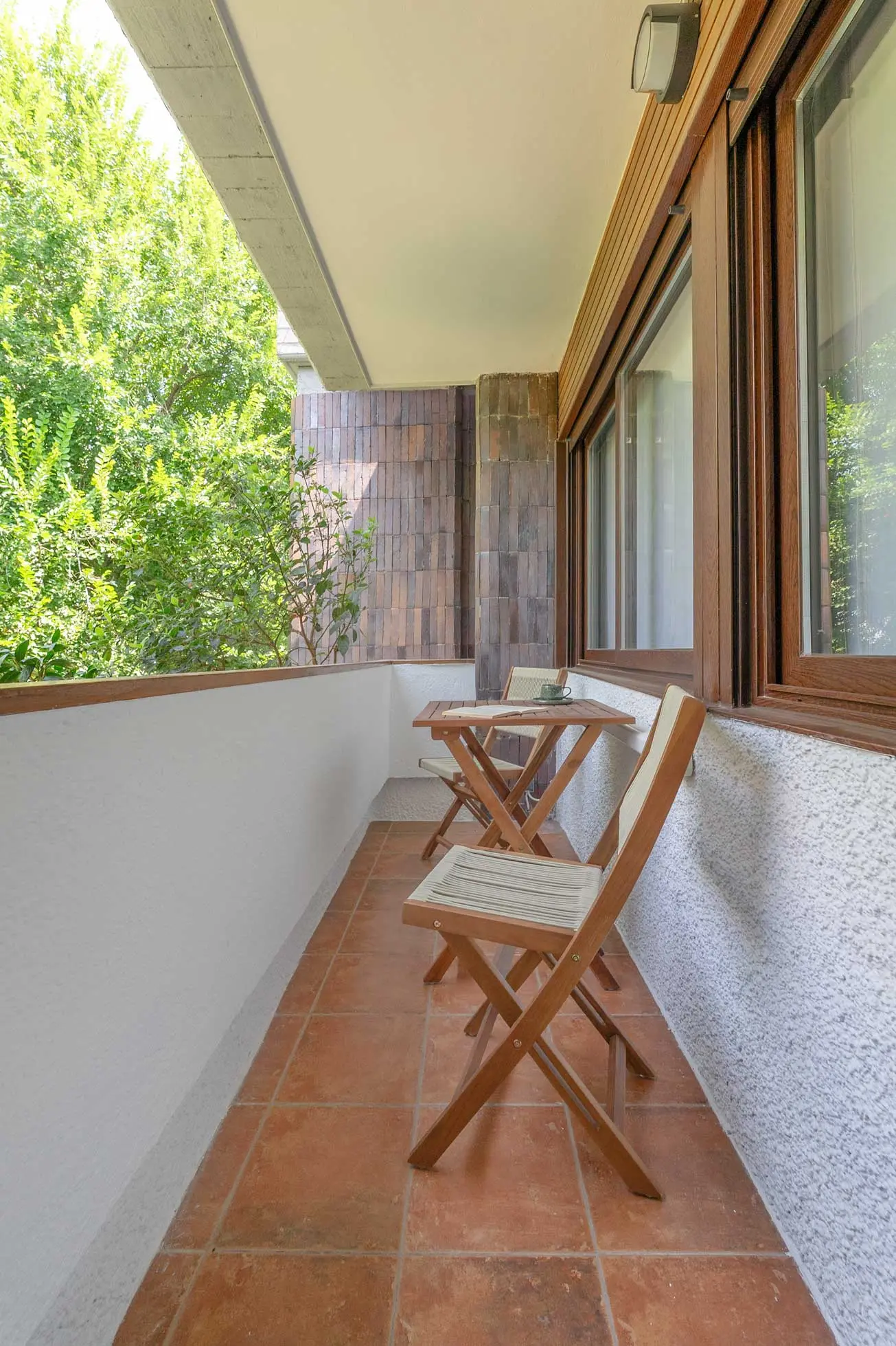 Terraza exterior alargada con suelo de baldosa de terracota, mesa y sillas plegables de madera y vistas a la arboleda