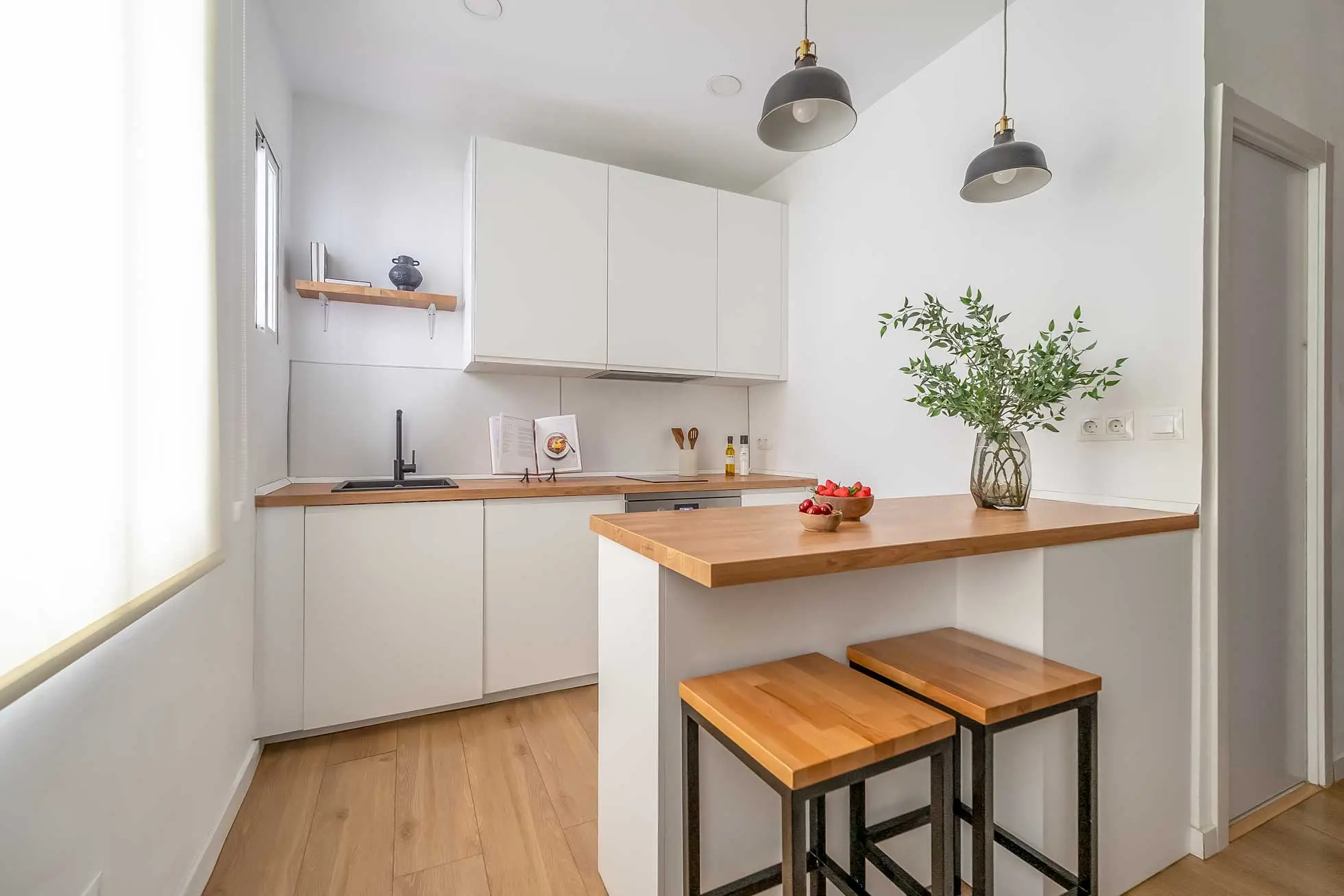 Vista cercana de la barra de la cocina con encimera de madera clara, taburetes industriales y jarrón decorativo con ramas verdes