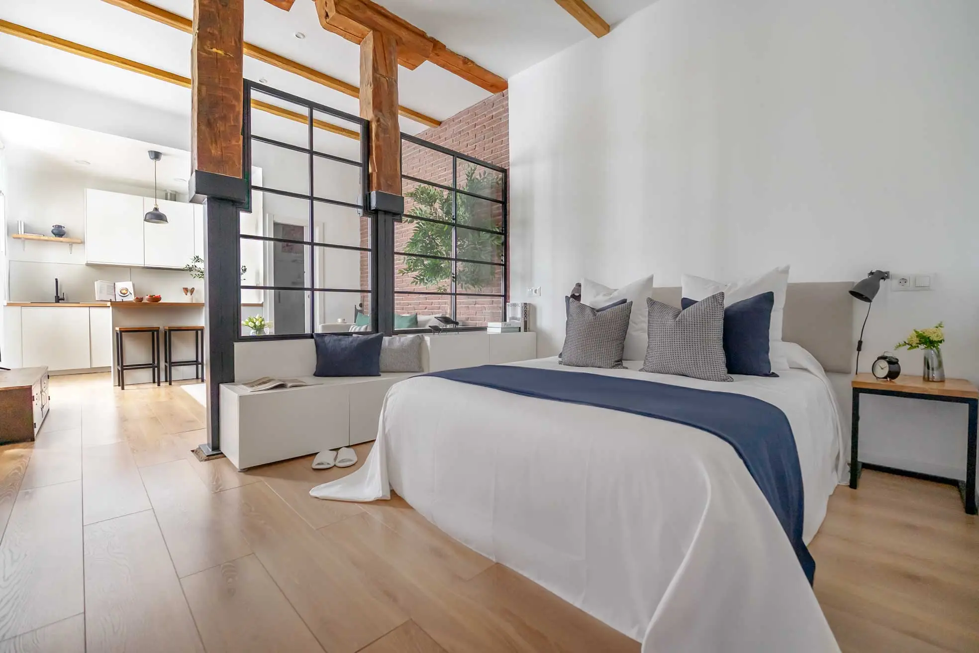 Perspectiva del dormitorio principal integrado visualmente con el salón mediante una gran cristalera industrial y vigas de madera vista