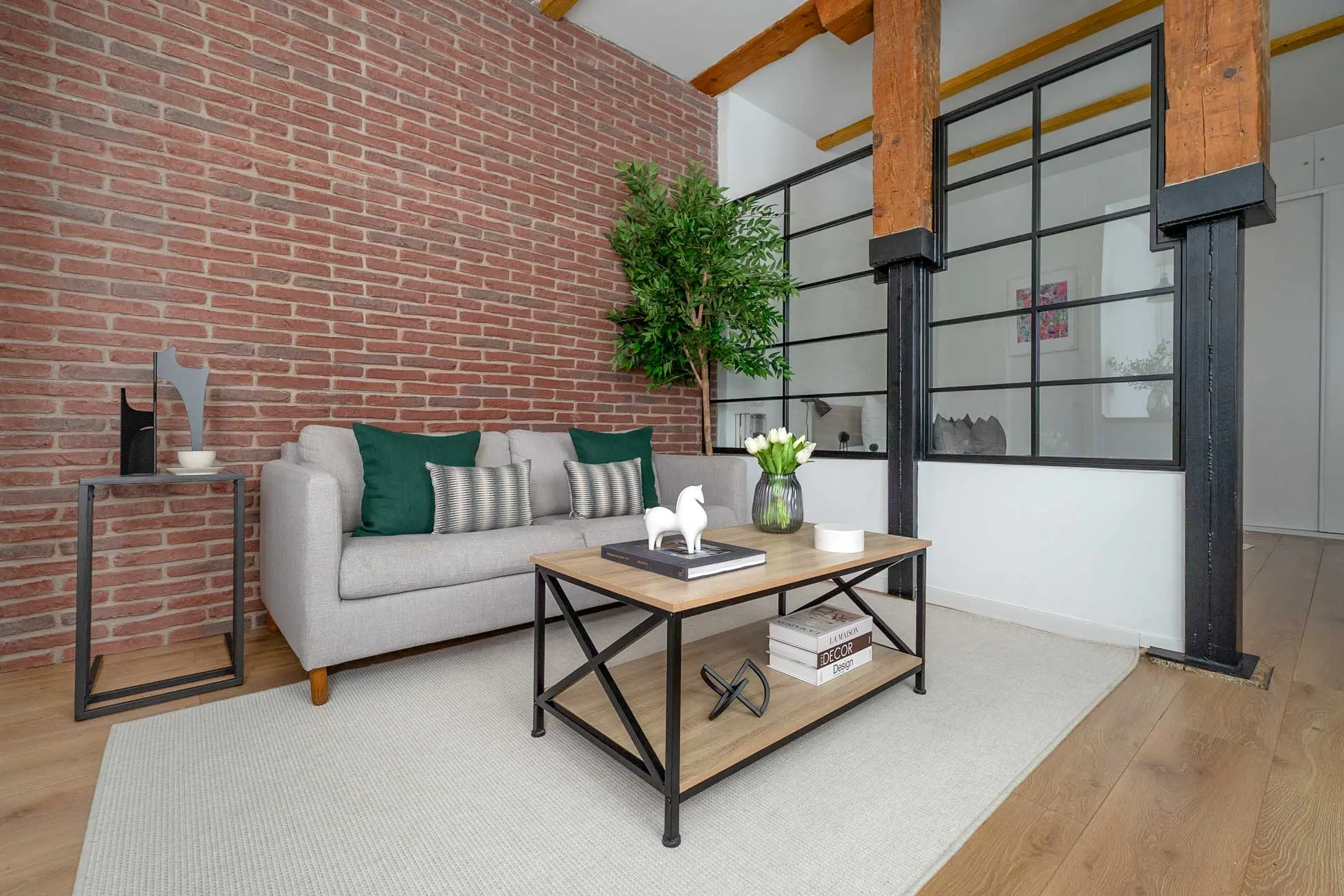 Zona de estar en el salón con sofá gris, cojines verde botella, mesa de centro de madera y una imponente pared de ladrillo antiguo