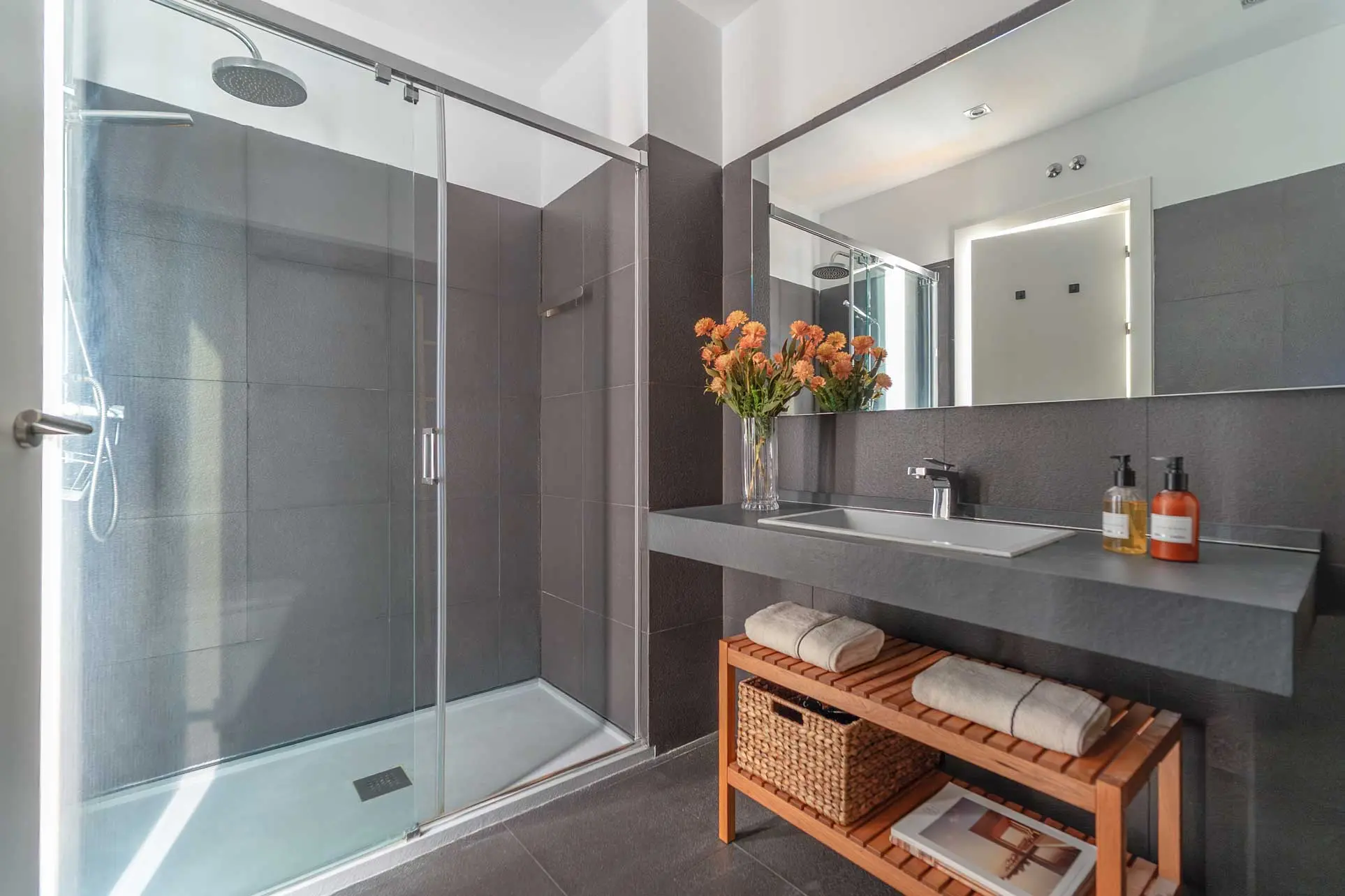 Segundo cuarto de baño con azulejos gris oscuro, plato de ducha con mampara de cristal y mueble de lavabo con estantes de madera
