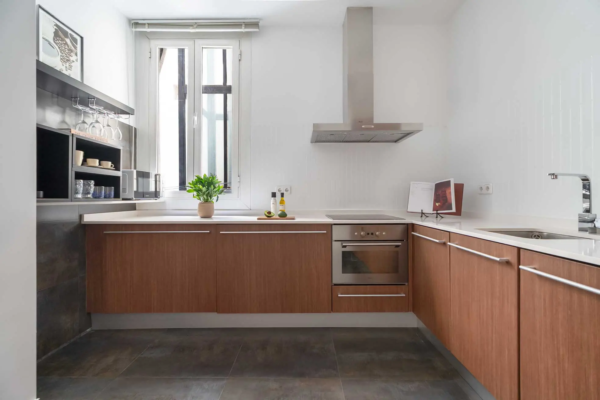 Perspectiva frontal de la cocina moderna con mobiliario en tonos madera, encimera blanca, rincón del café integrado y ventana al fondo en el apartamento Fuencarral