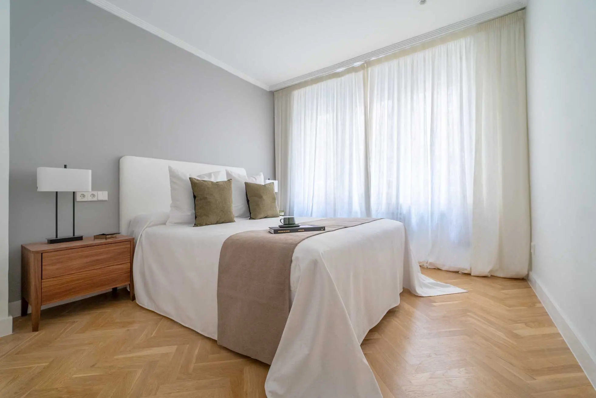 Dormitorio de invitados con cama de matrimonio vestida de blanco, camino beige, cojines verde oliva y suelo en espiga