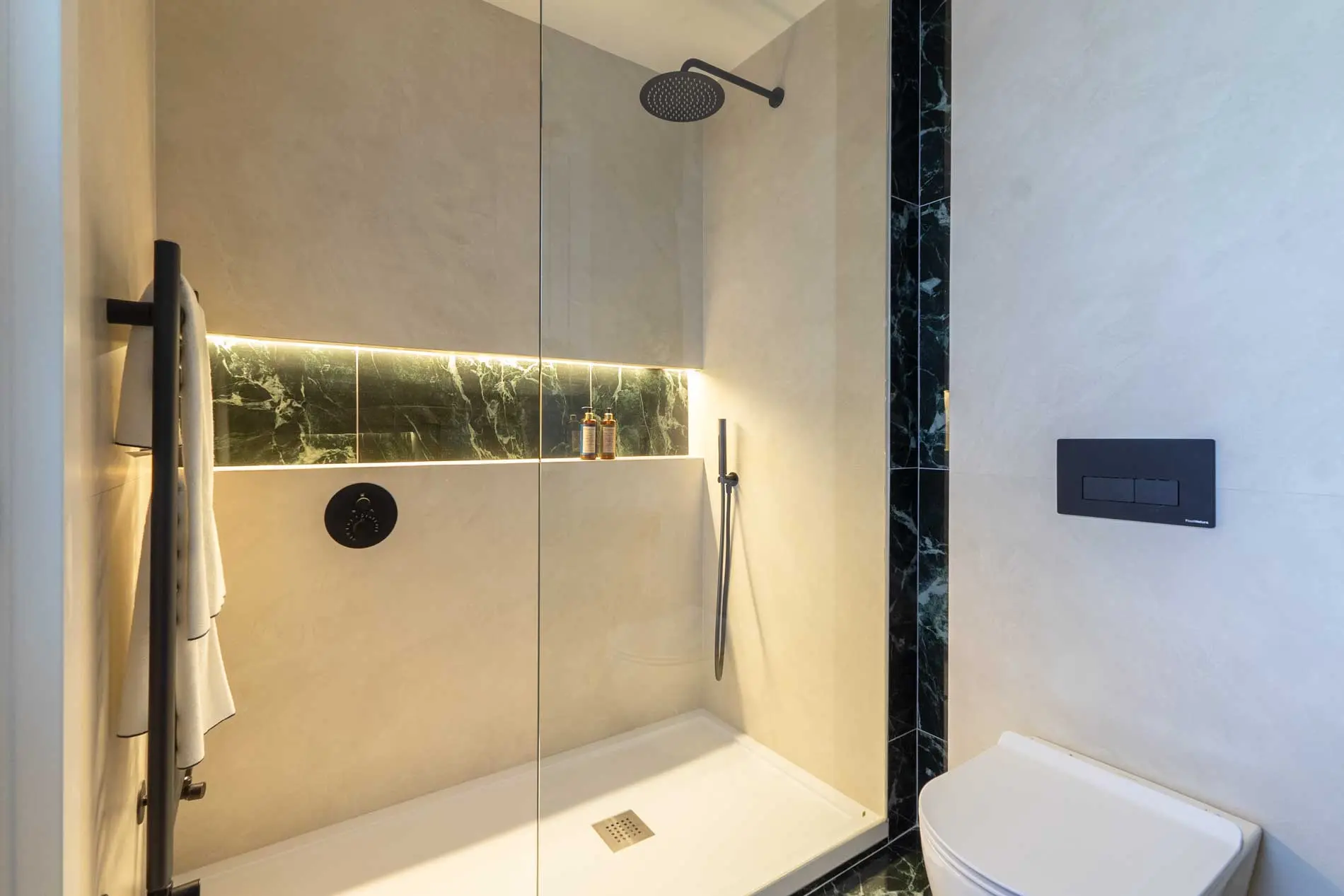 Cuarto de baño de lujo con ducha italiana, revestimiento cerámico beige y hornacina iluminada con mármol veteado verde oscuro