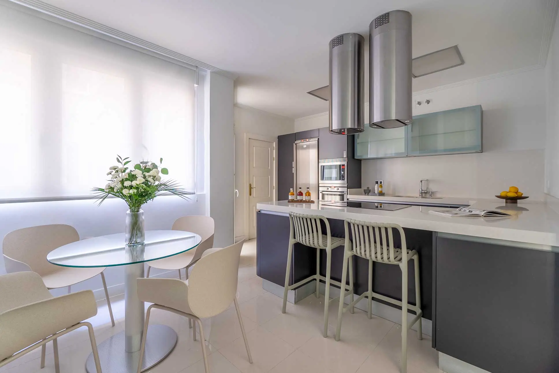 Cocina de diseño contemporáneo con isla central, taburetes altos en tonos claros, campanas extractoras cilíndricas de acero y mesa de comedor de cristal integrada
