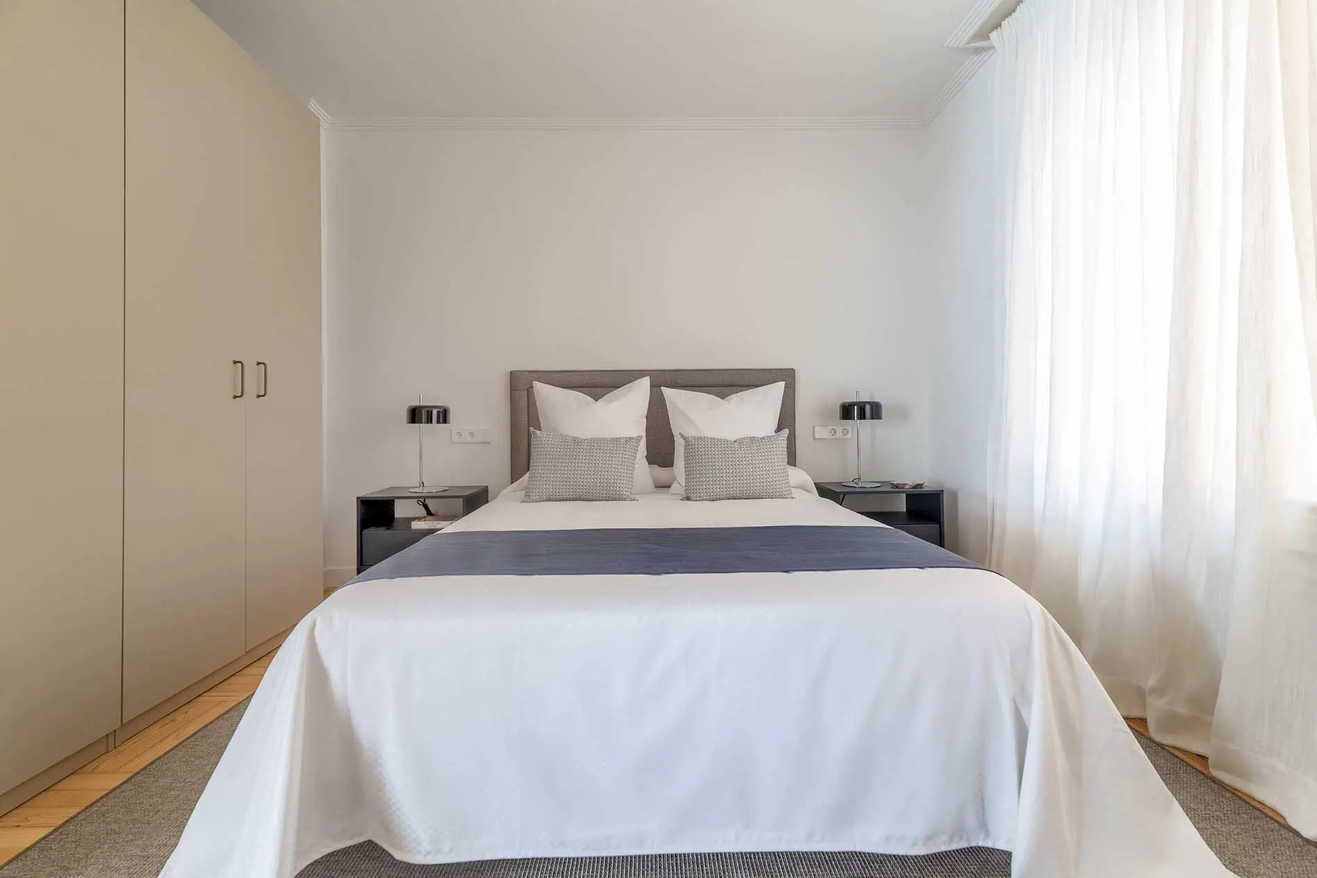 Perspectiva frontal del segundo dormitorio con cama doble vestida minuciosamente con textiles blancos y camino azul marino junto a armarios modulares nudes