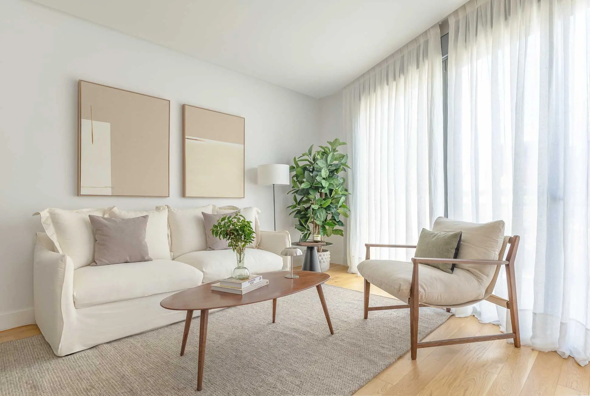 Salón luminoso con sofá blanco de fundas, cojines en tonos malva, mesa de centro ovalada de madera, butaca de estructura de madera y grandes ventanales con visillos.