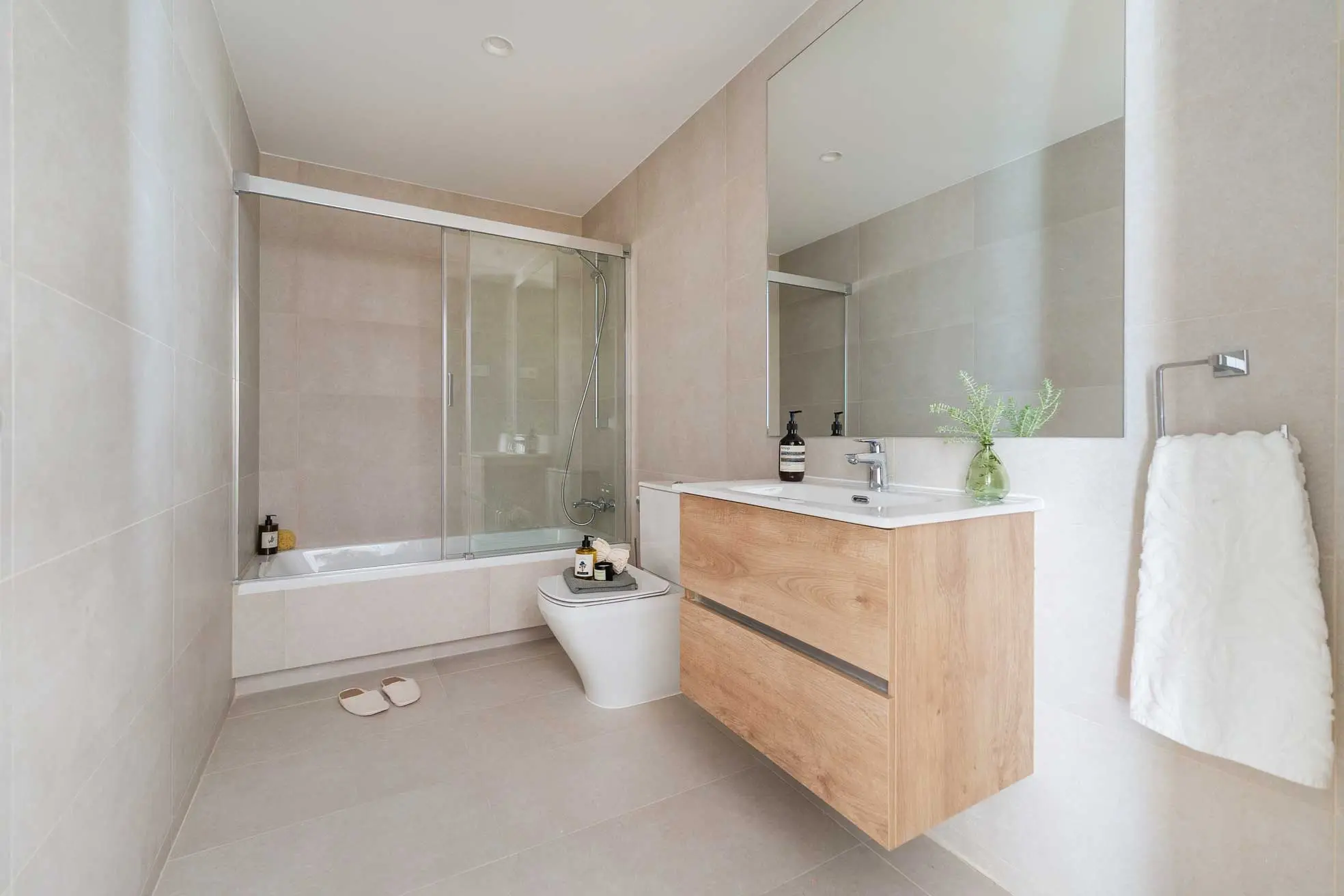 Baño amplio revestido en gres beige, con mueble de lavabo de madera suspendido, gran espejo, inodoro y bañera con mampara de cristal corredera.